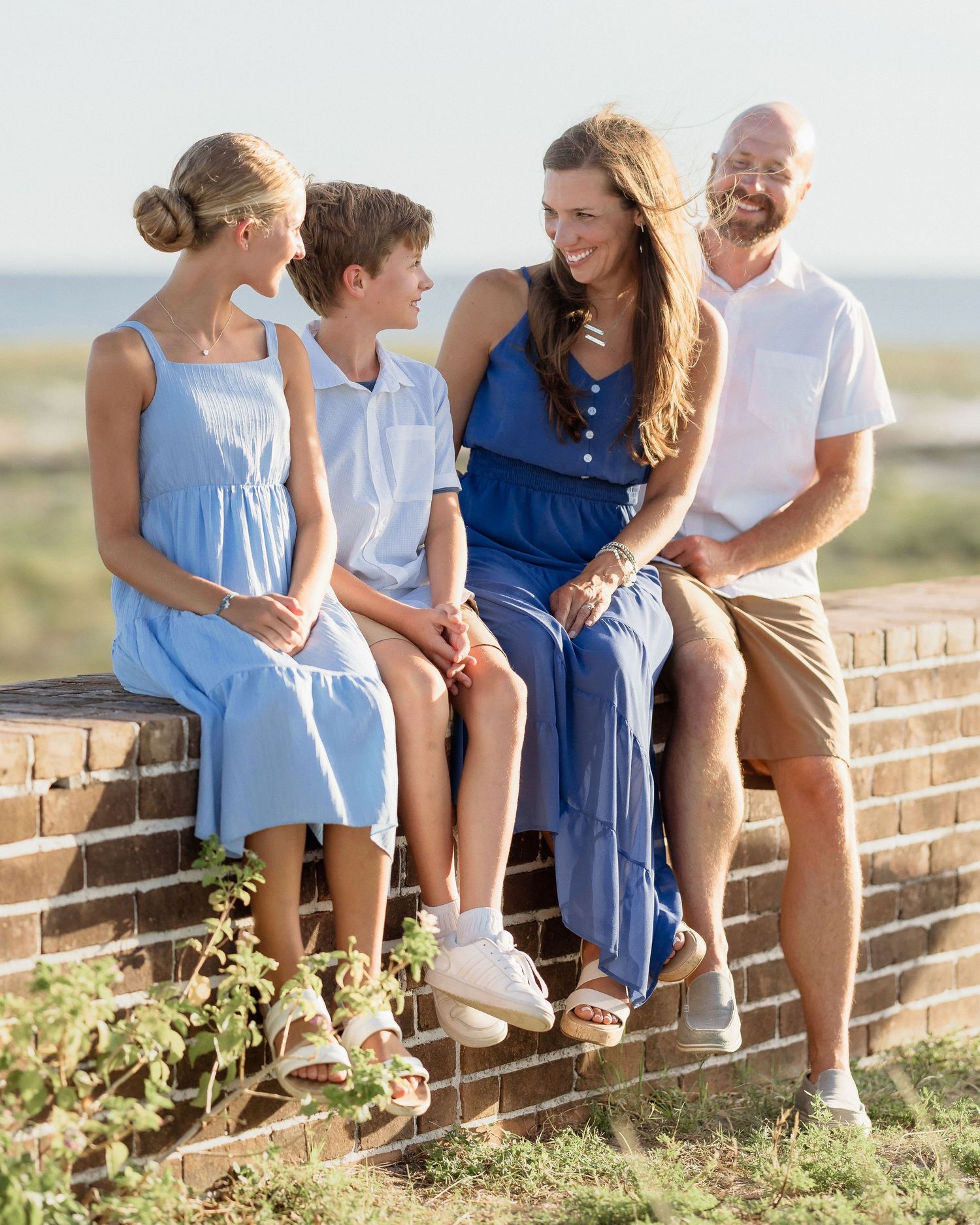 A happy family of four sits on a brick wall near a beach, dressed in coordinating blue and white outfits.