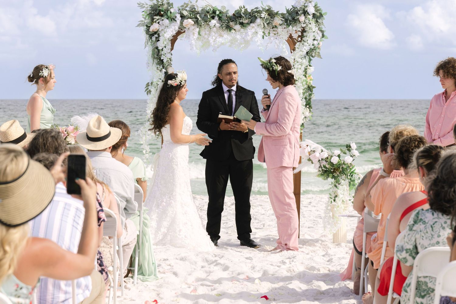 Beach wedding ceremony with bride and groom at floral arch, officiant in pink, guests seated on sandy shore.