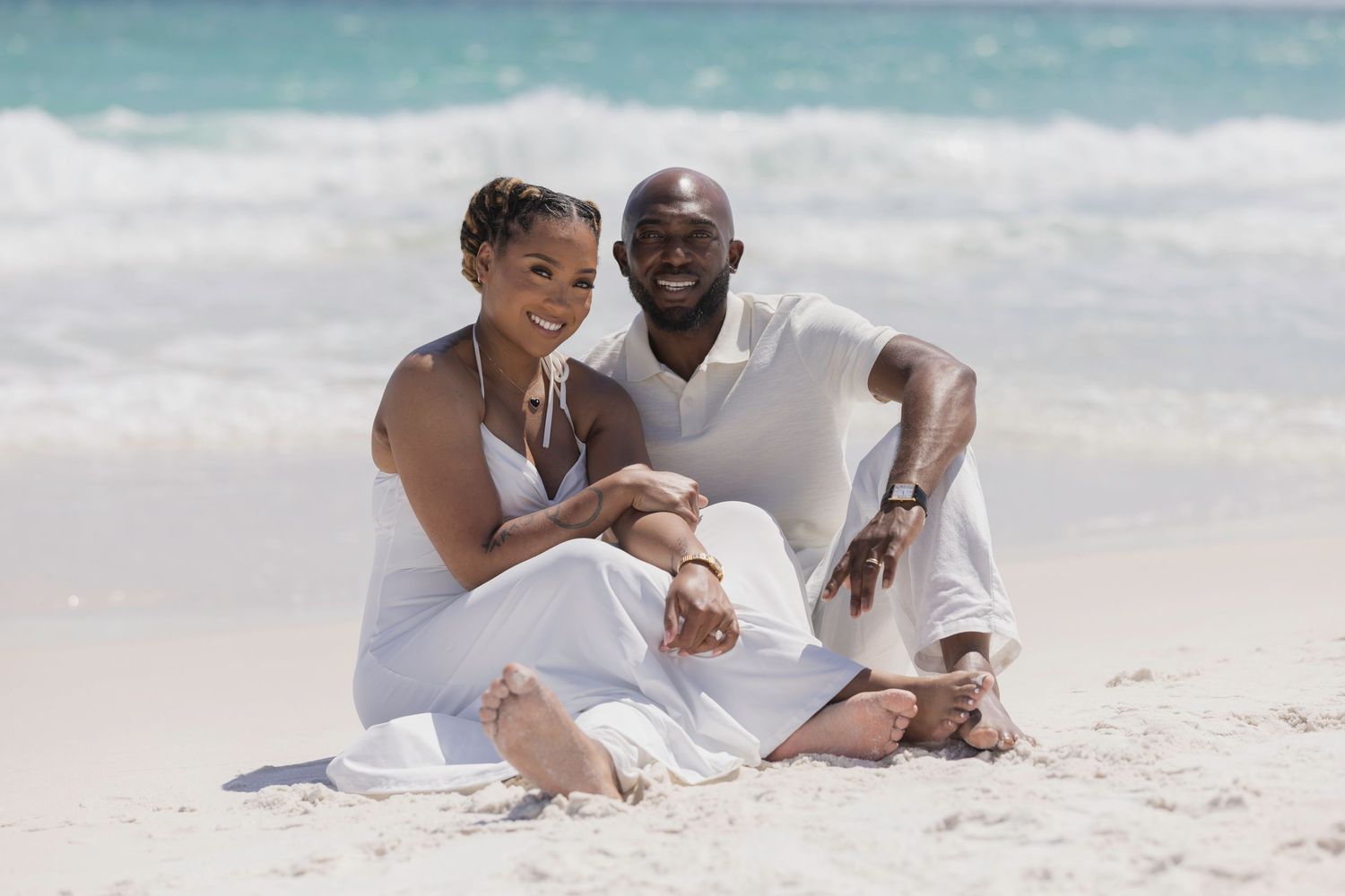 Happy couple in white outfits sitting together on a sandy beach with turquoise ocean waves in the background.
