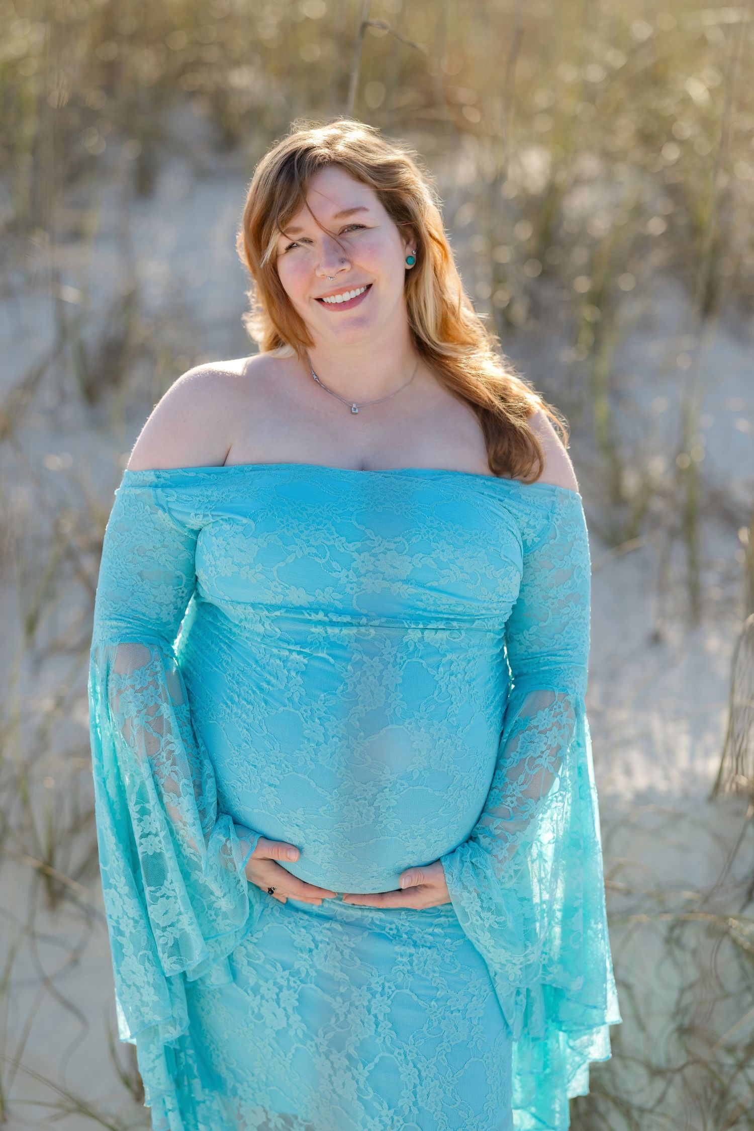 Smiling pregnant woman in teal lace off-shoulder maternity gown holding belly on sandy beach dunes.