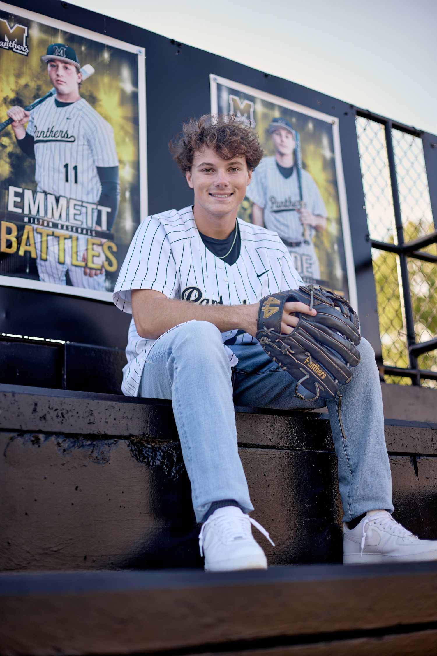 Smiling teen in Panthers baseball jersey sits in dugout beside Emmett Battles player poster holding glove.