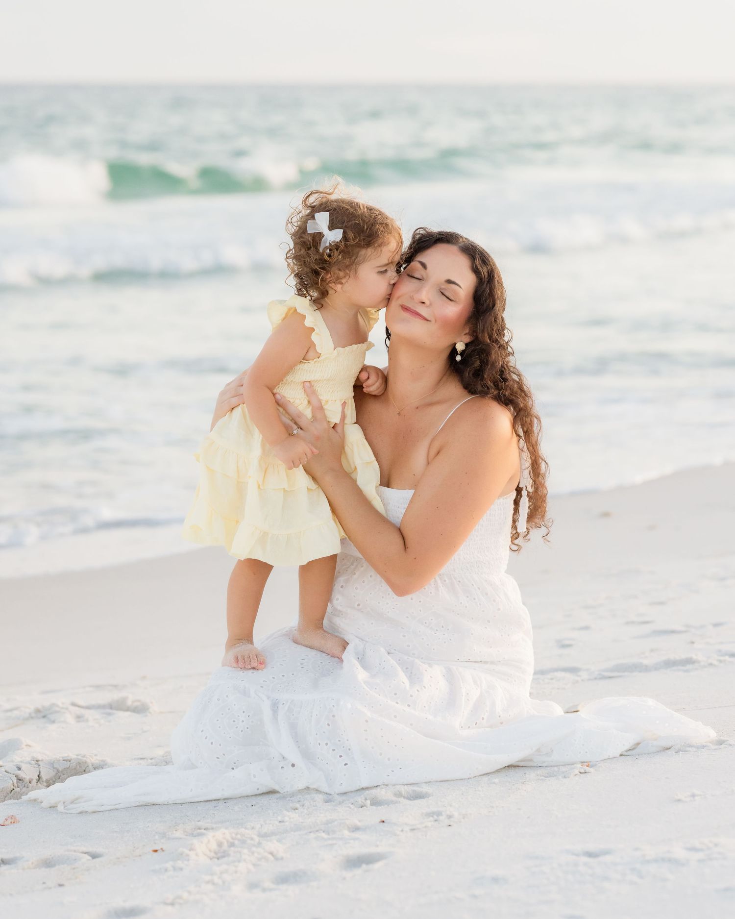 Mother in white dress sitting on beach sand holding young daughter in yellow dress sharing a kiss.