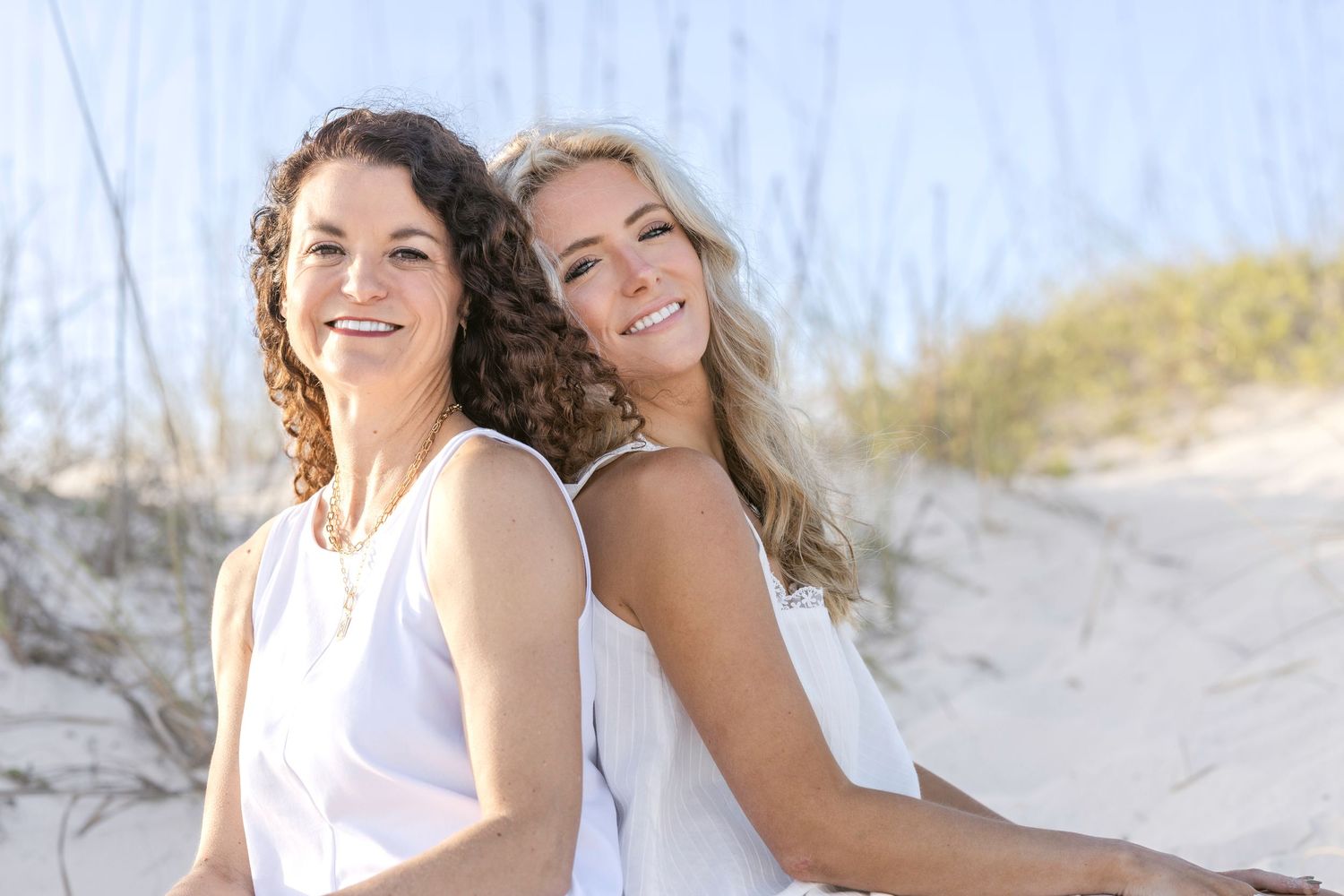Two smiling women in white outfits sitting back-to-back on sandy beach dunes on a sunny day.