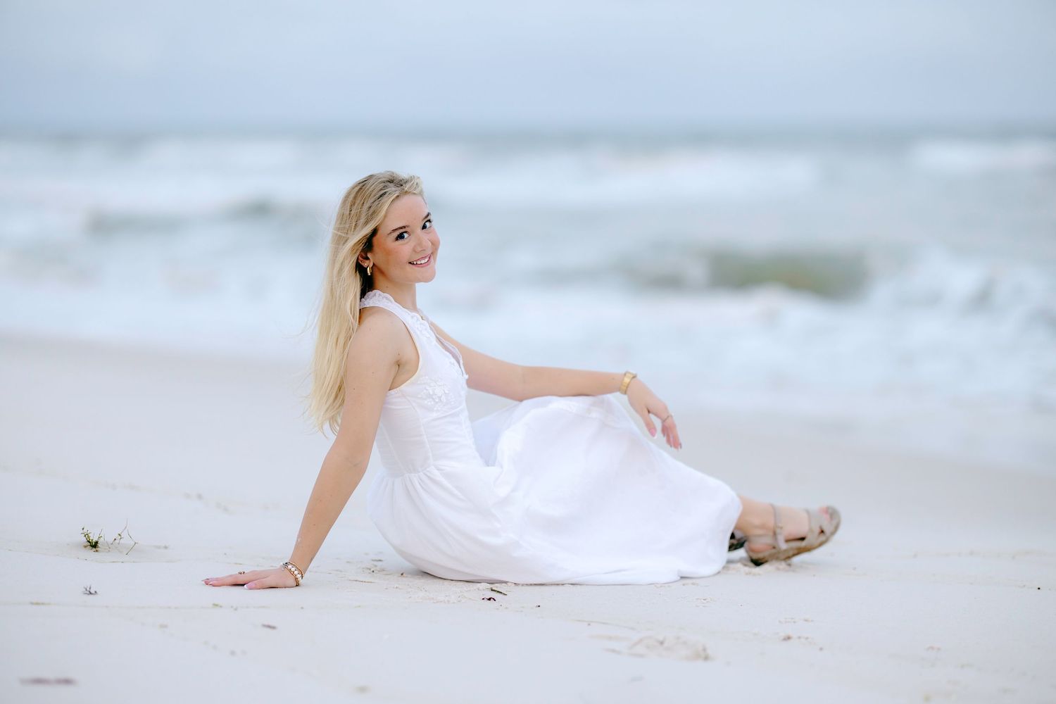Woman in flowing white dress sits gracefully on sandy beach at sunset.