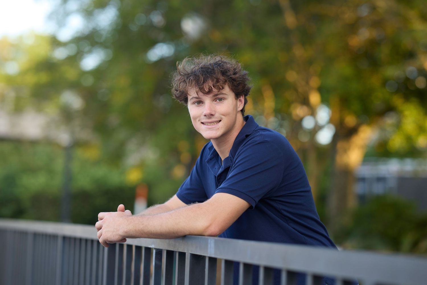 Casual outdoor portrait of young person in navy polo leaning on railing.