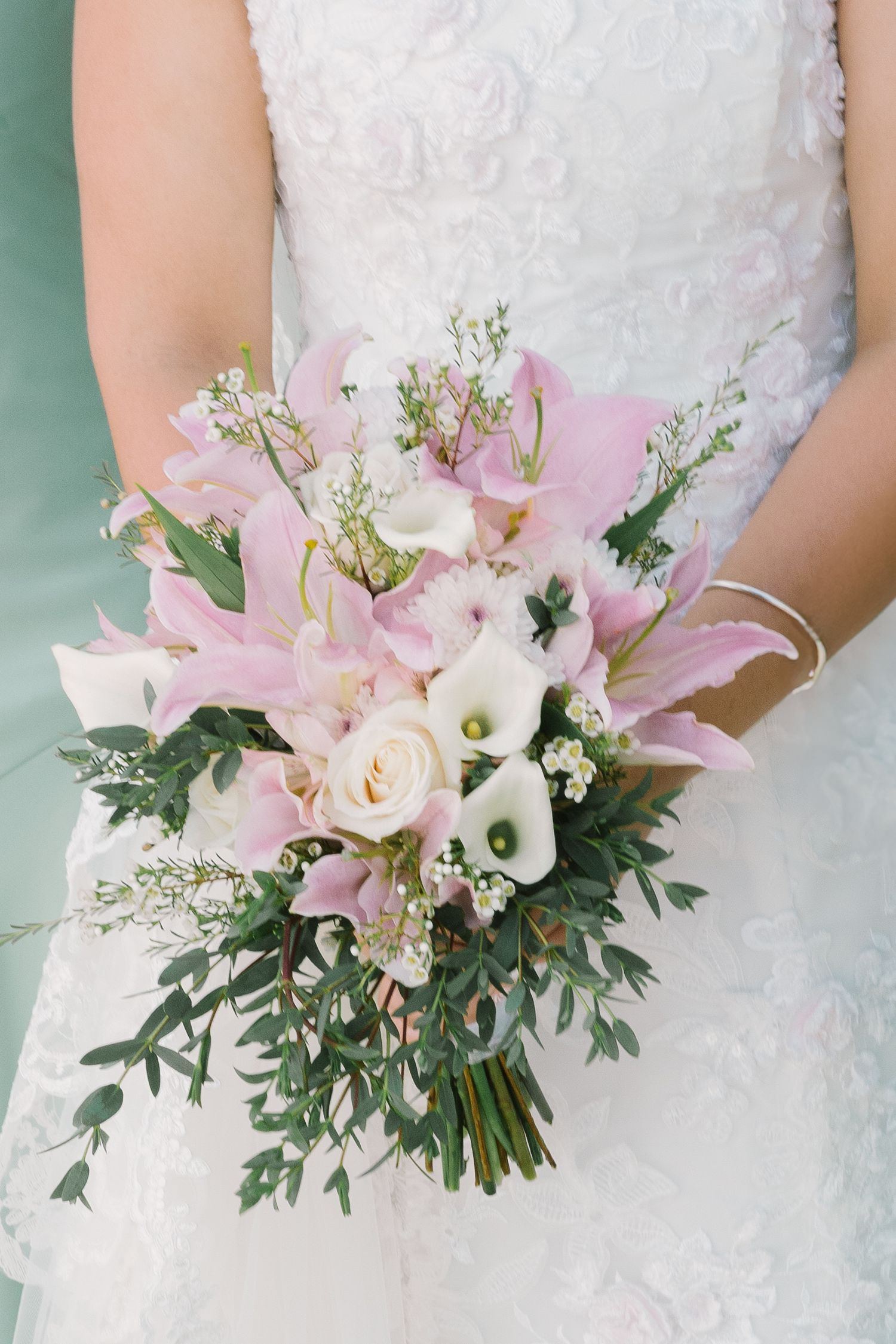 Elegant bridal bouquet featuring pink roses, calla lilies and eucalyptus against white lace dress.