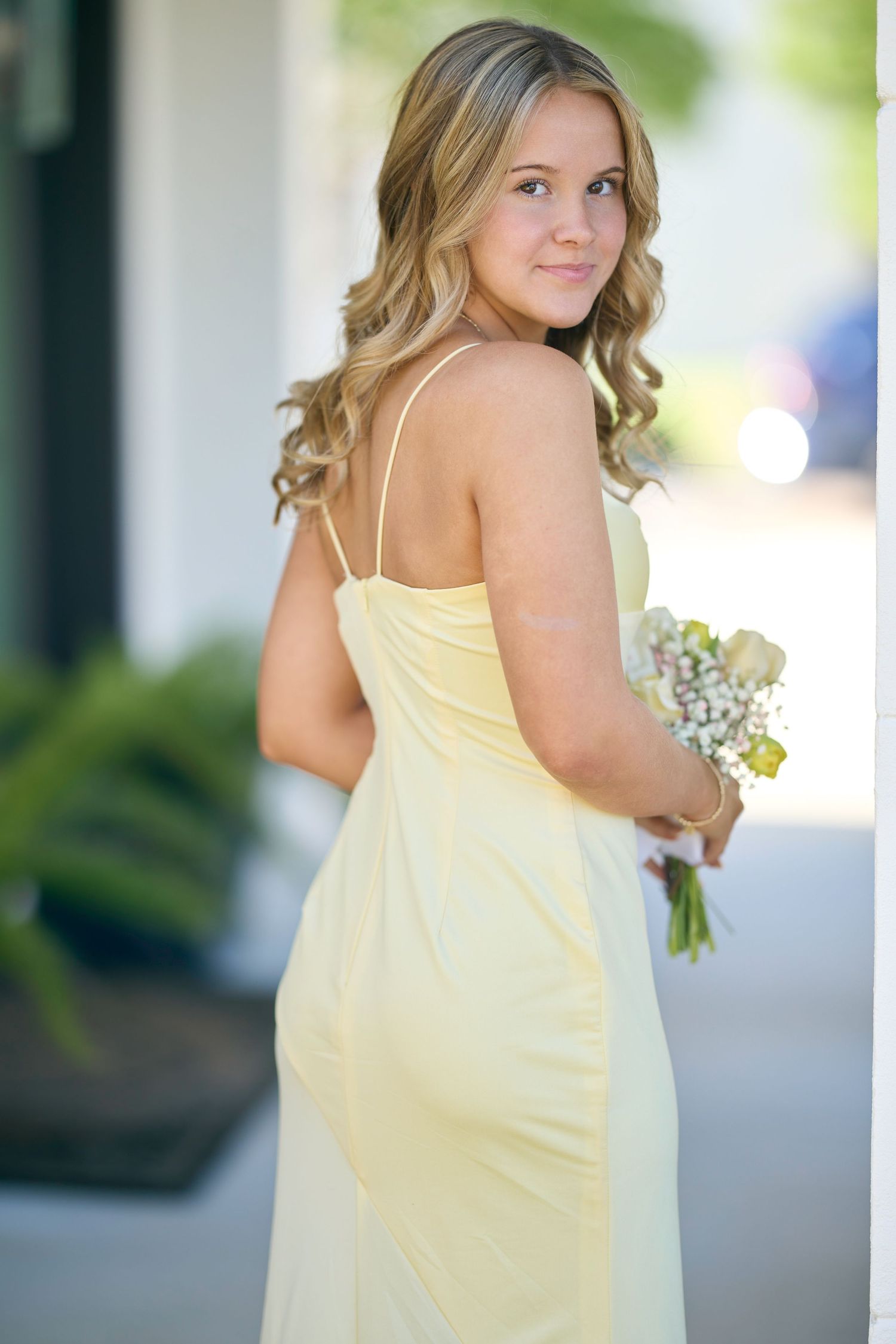 Young woman in yellow spaghetti strap gown holding bouquet of white flowers, posing outdoors.