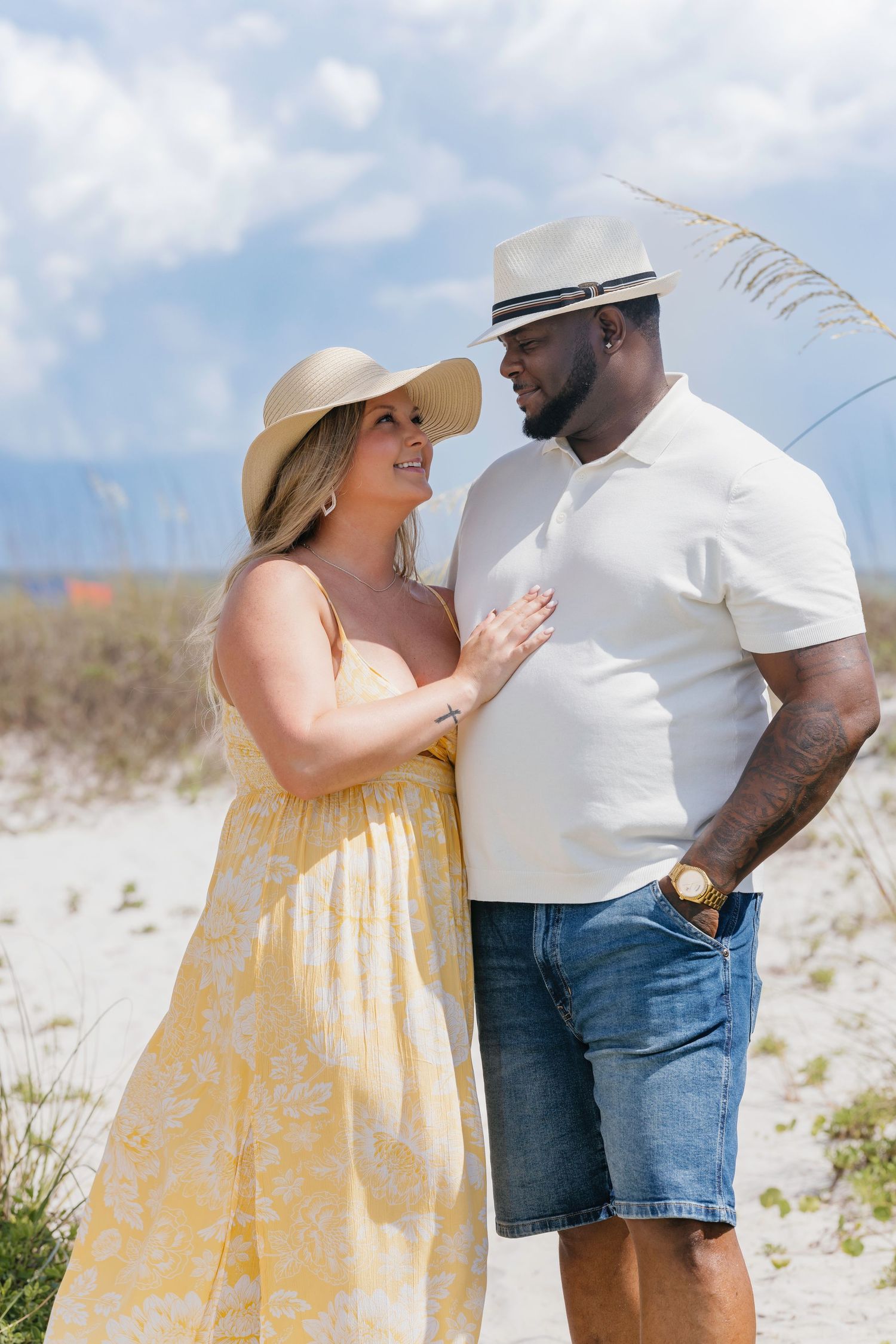 Couple in beach attire gazing at each other on sandy dunes, woman in yellow dress and sun hat, man in white polo.