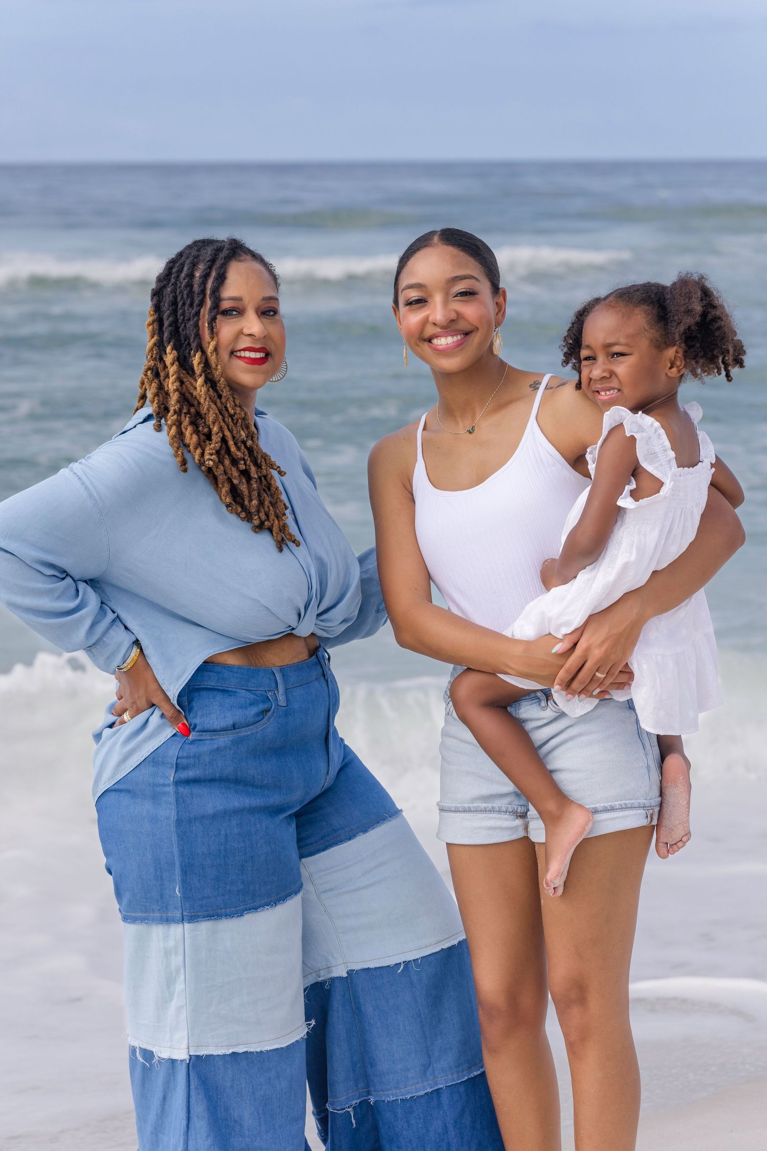 Three generations of Black women pose together on a beach, with waves in the background.
