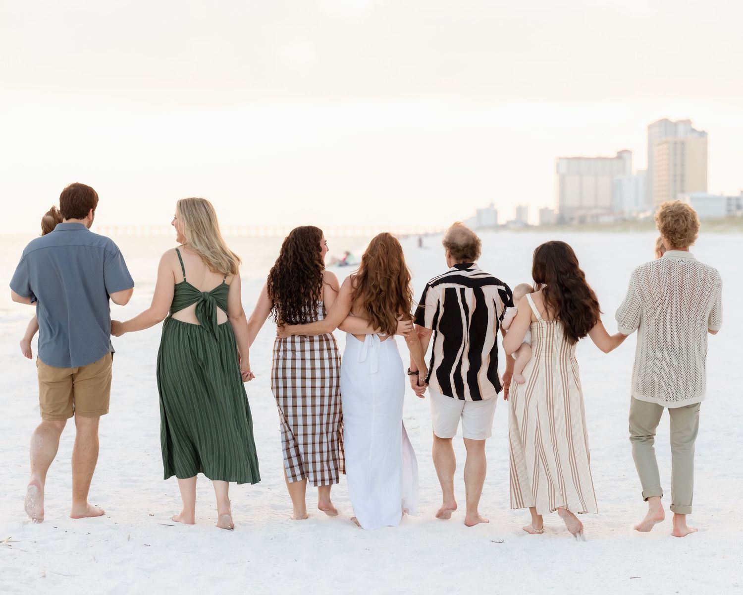 Group of seven friends holding hands on a white sand beach, viewed from behind, wearing summer outfits at sunset.