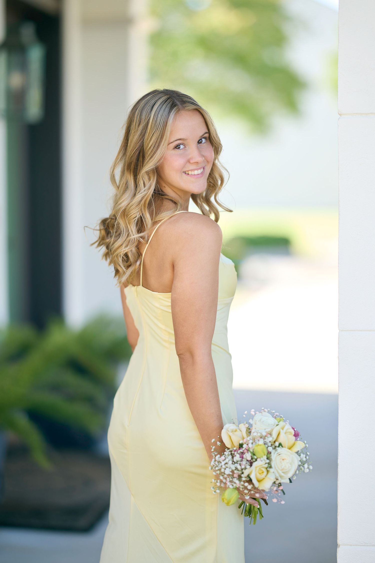 A person in a pale yellow dress poses with a bouquet of white flowers on a porch with columns.