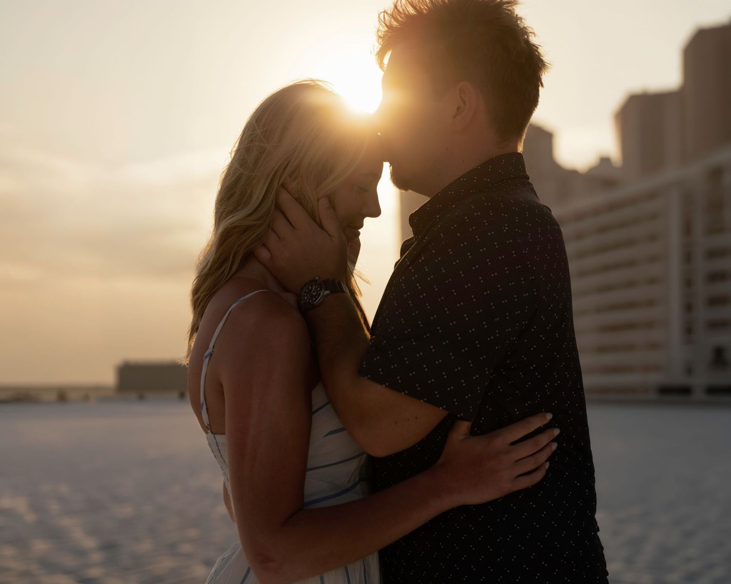 Couple embracing at sunset on a beach with golden backlight and city buildings in the background.