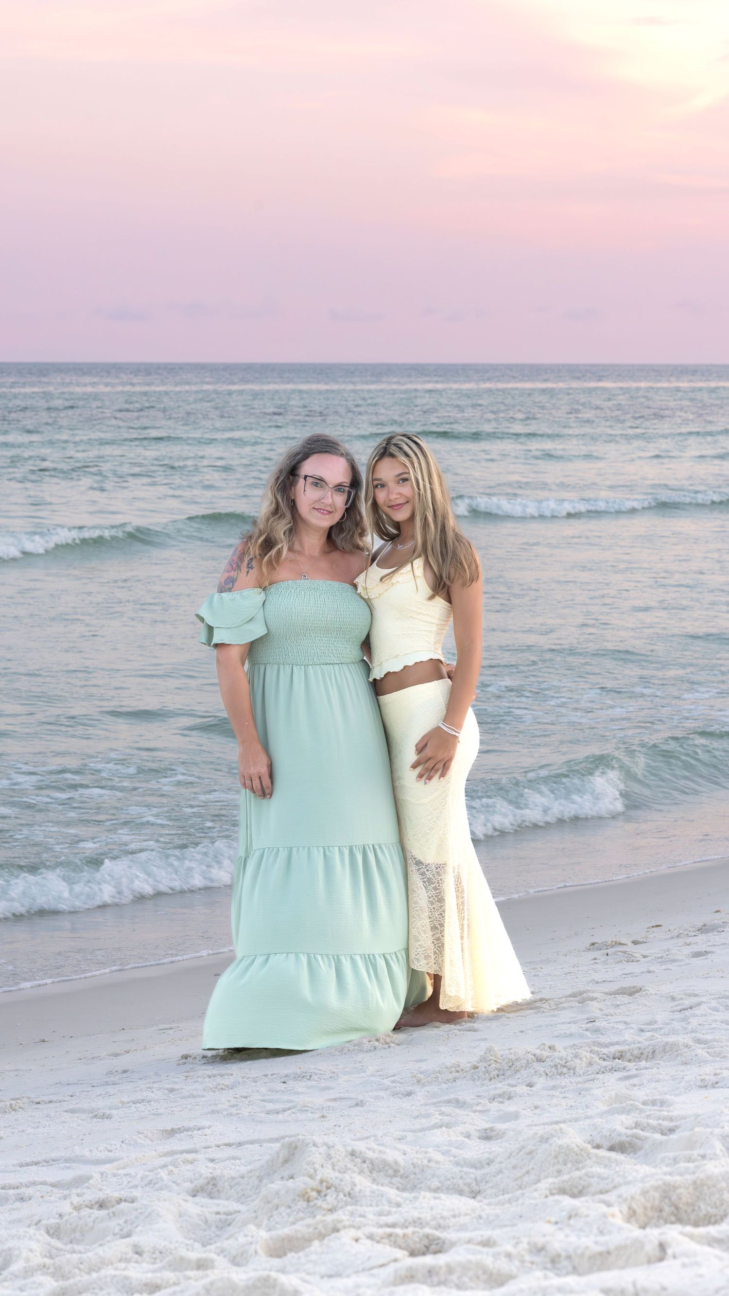 Two friends in matching dresses pose together on the beach at sunset with gentle waves rolling in behind them.