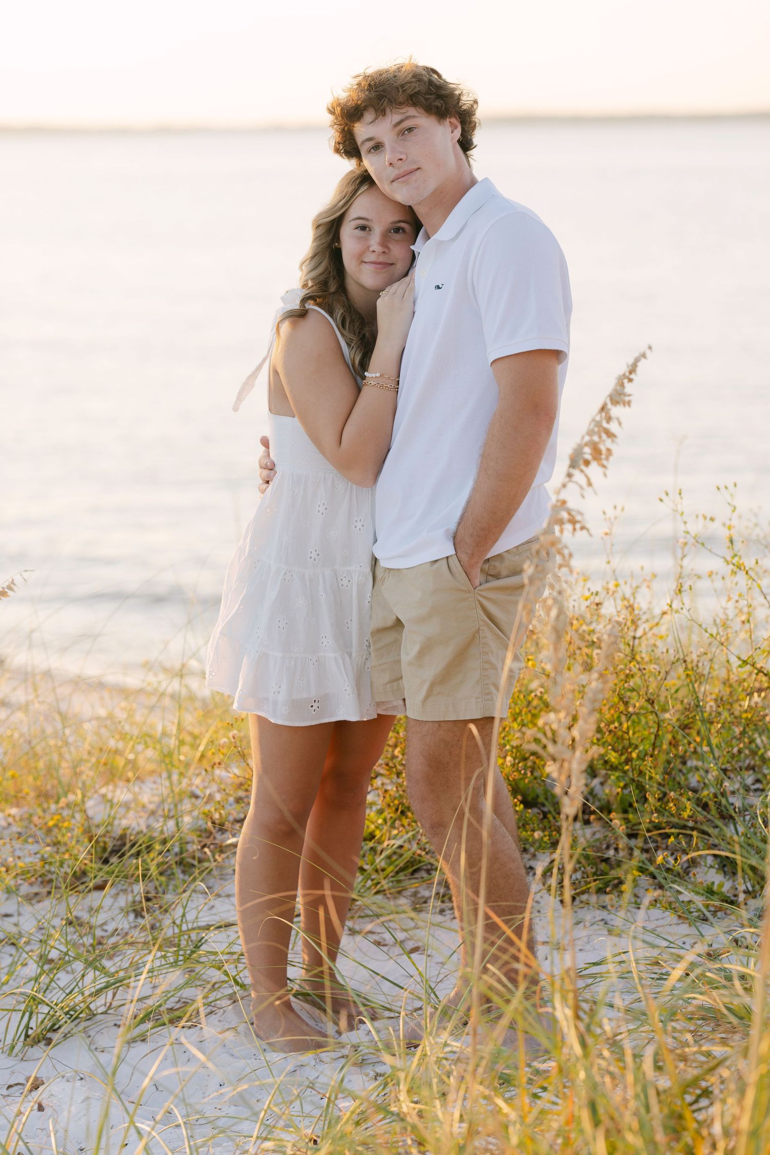 A couple embraces on a beach at sunset among sea oats and sand dunes in casual summer attire.