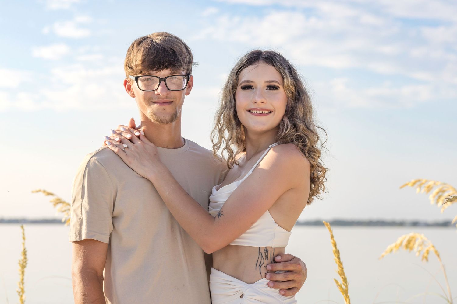 Happy couple embraces near water at sunset with sea oats in foreground and soft sky behind them.