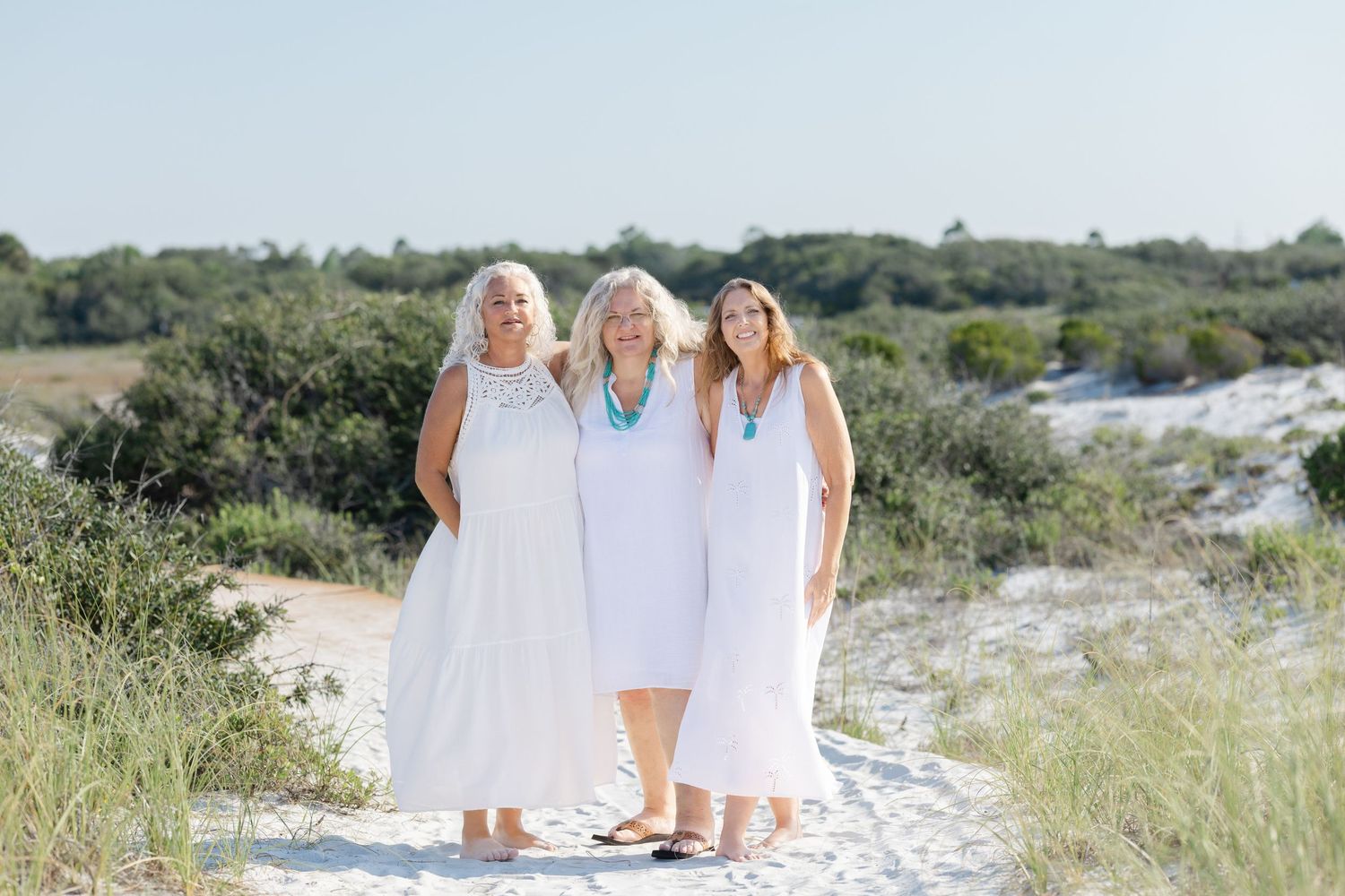 Three women wearing white dresses pose together on a sandy beach path with green vegetation in the background.