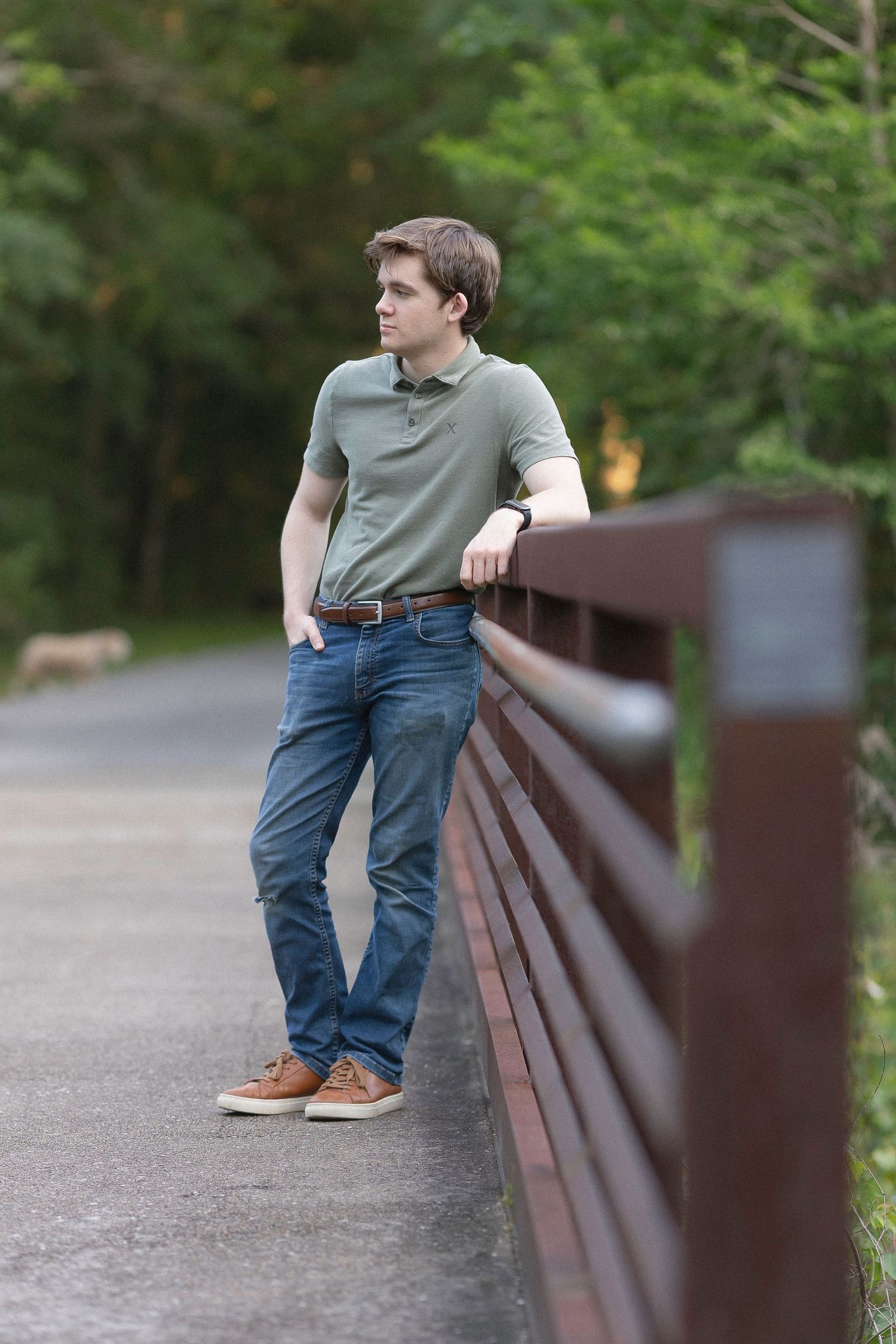 Person wearing jeans and gray t-shirt leans casually against wooden railing on outdoor path surrounded by greenery.