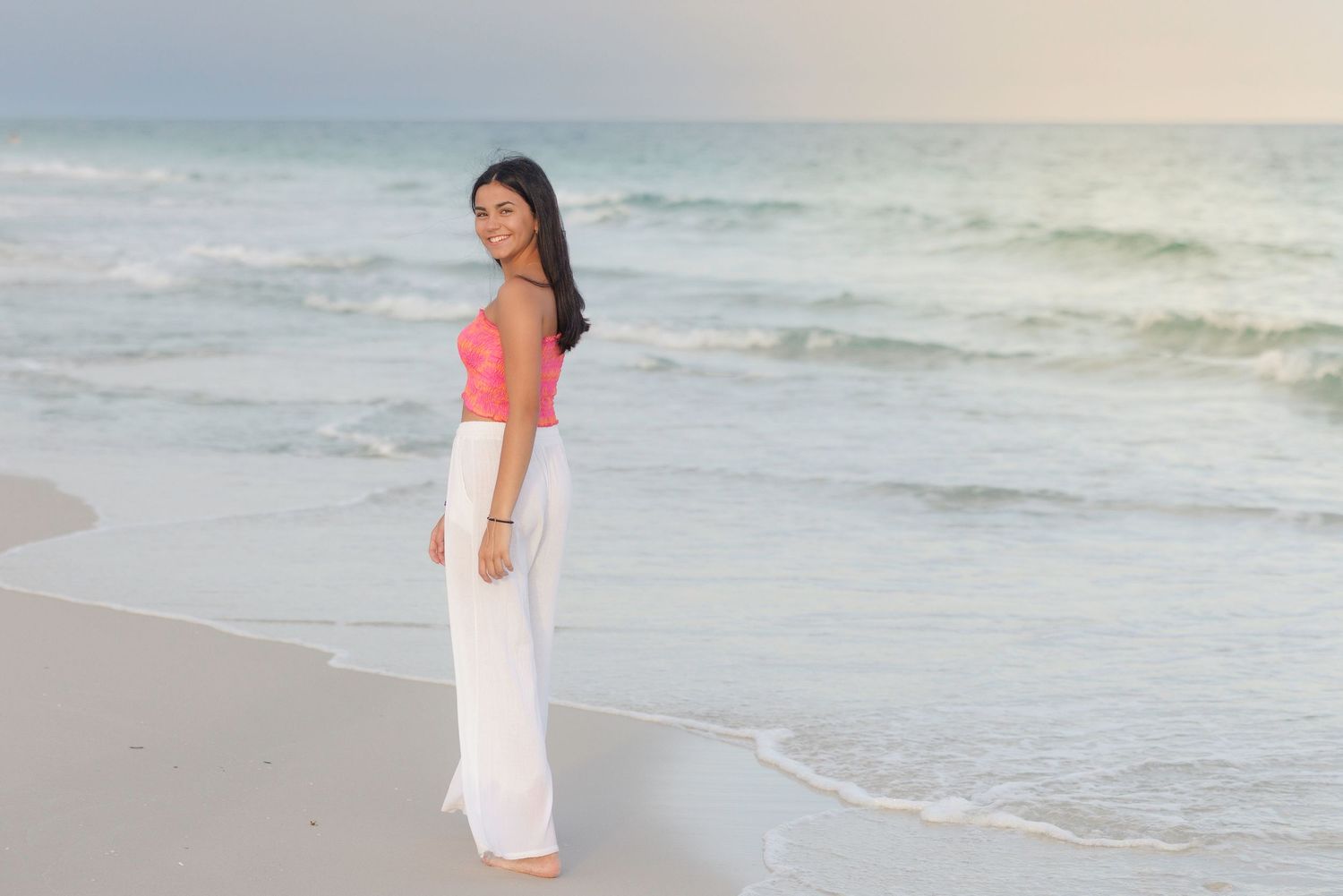 Casual beach portraits with white flowing pants and coral top along the shoreline at sunset.