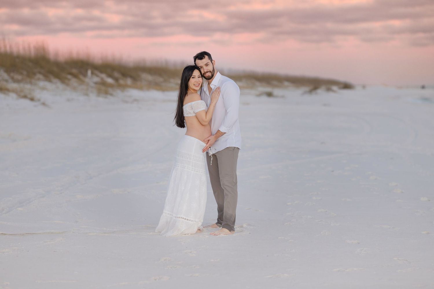 A couple in wedding attire embraces on a white sandy beach during a romantic sunset photoshoot.