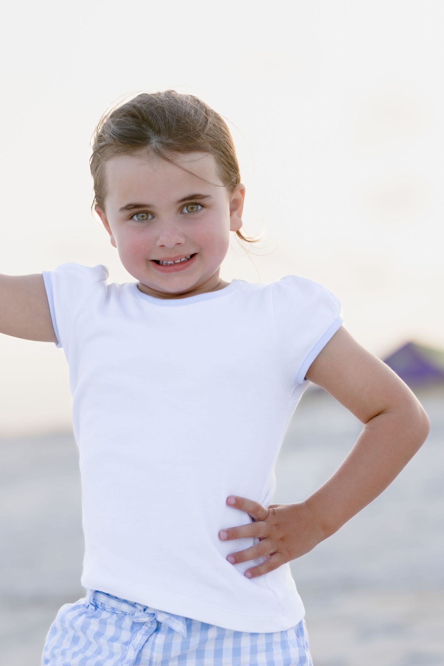 Young girl in white t-shirt and blue checkered shorts posing confidently on a beach with hand on hip.
