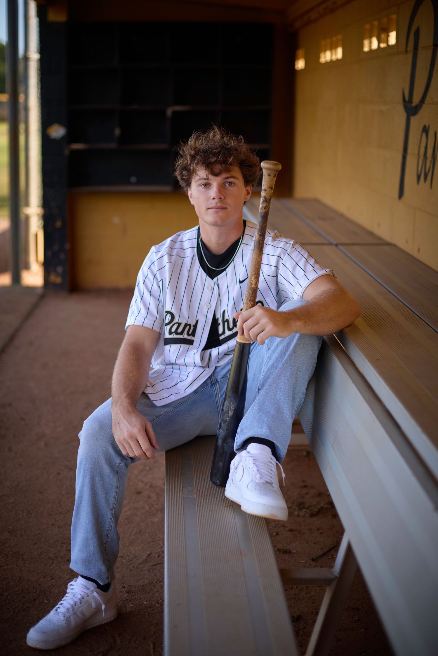Young baseball player in Panthers uniform sits in dugout holding bat, wearing jeans and white sneakers.