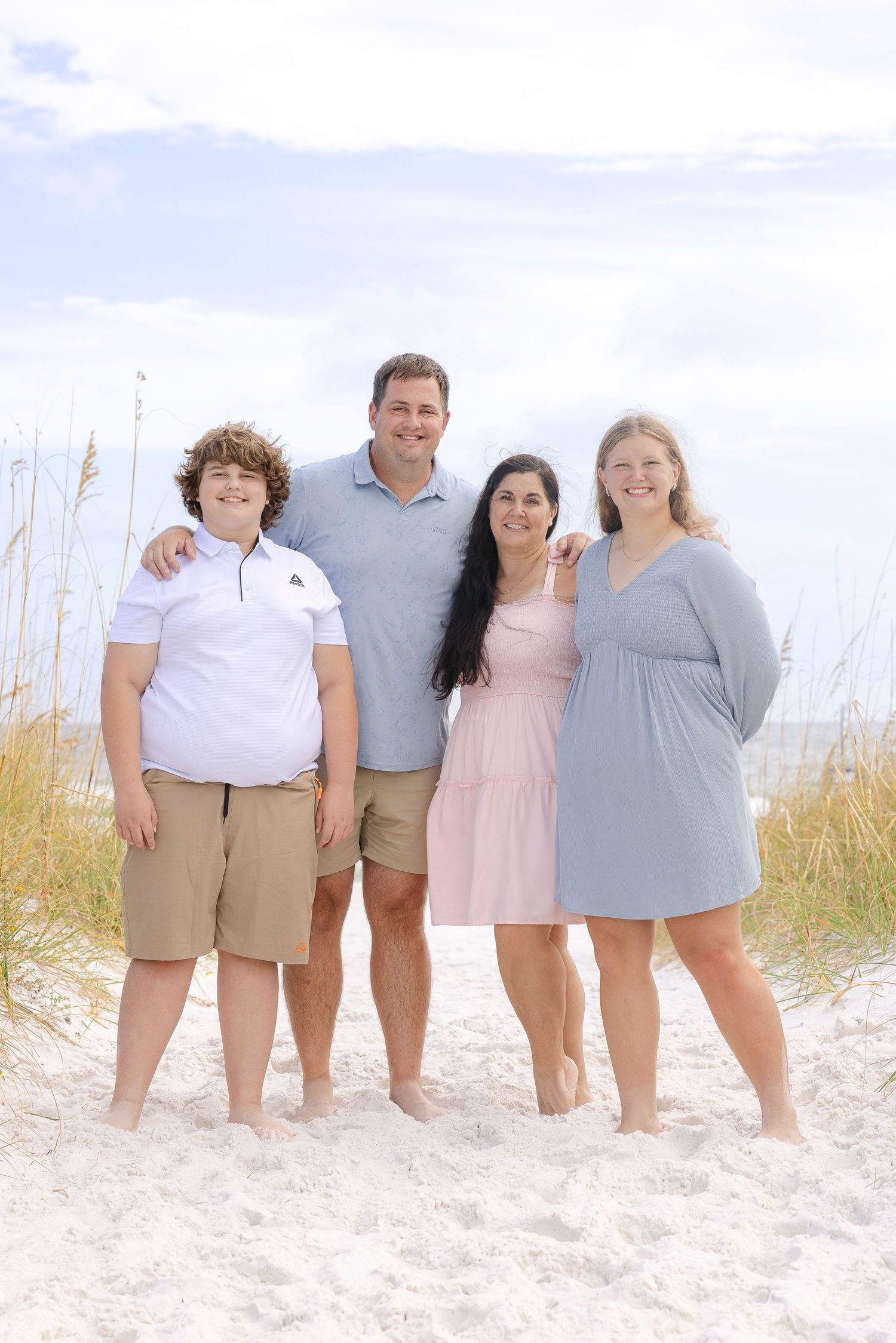 Family members standing together on a sandy beach with sea grass and blue skies in the background.
