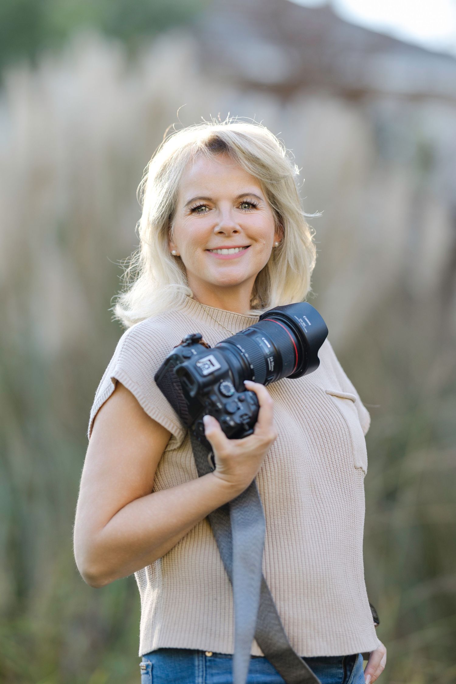 A photographer in a beige top holds a professional camera with telephoto lens outdoors.