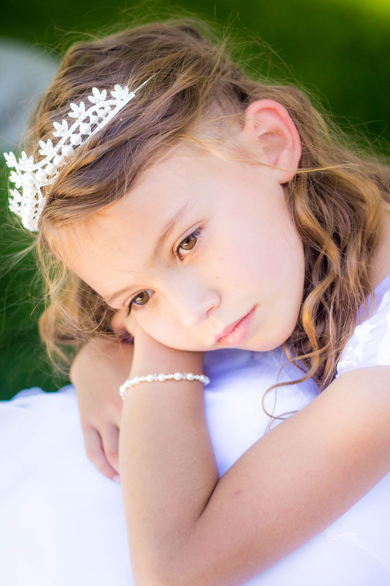 Child in white dress with floral hair accessories rests on white surface outdoors.