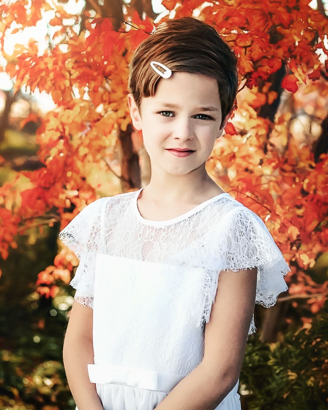 Child in white lace dress poses against vibrant red autumn foliage.