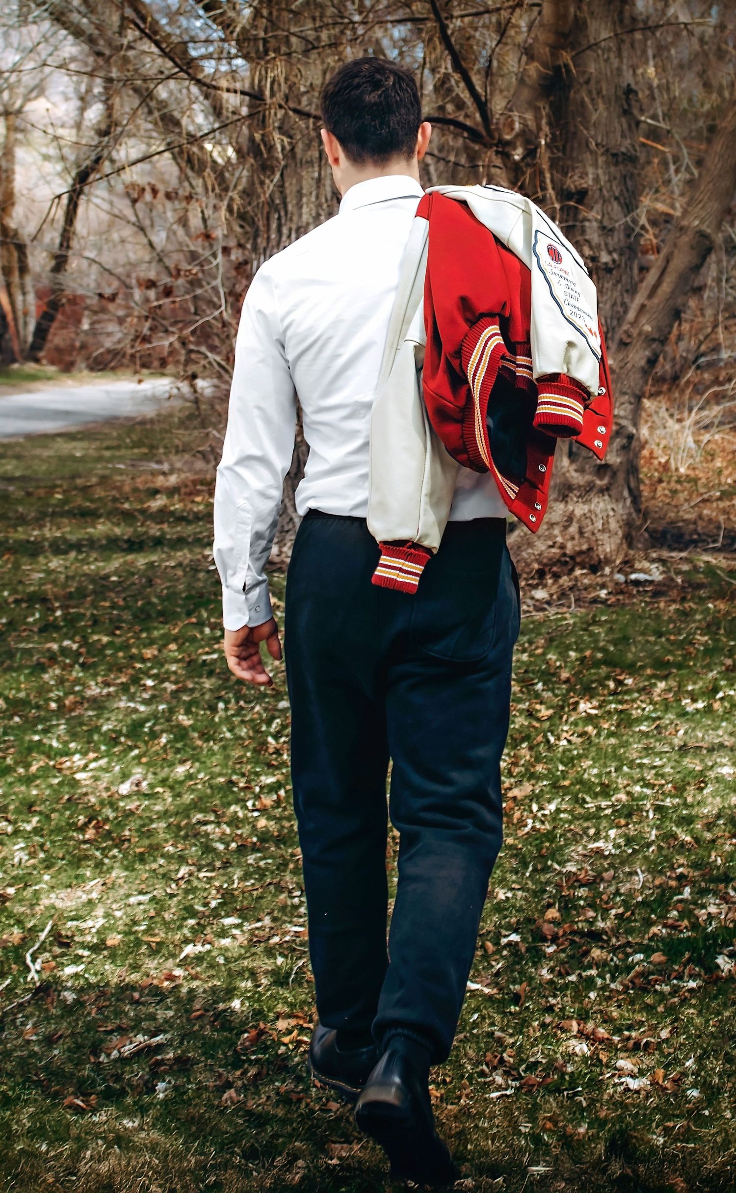 Back view of someone in formal attire carrying a red and white varsity jacket through a park.
