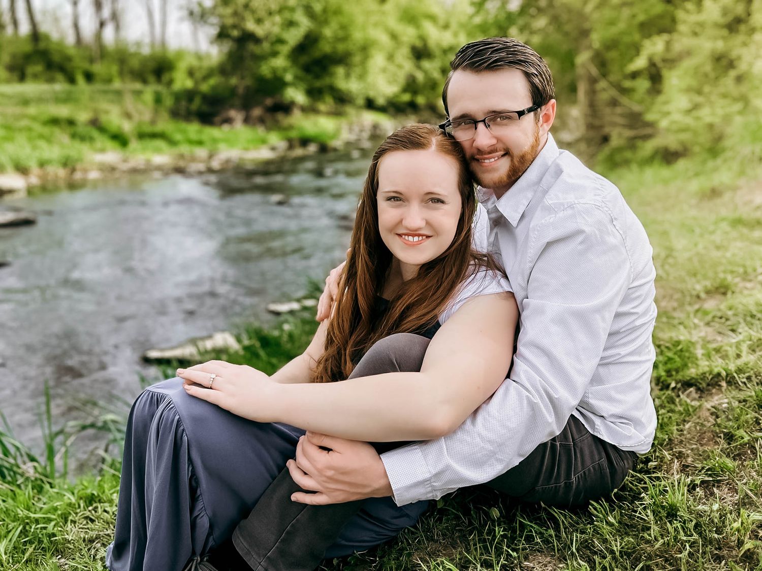 Couple sitting together on grassy riverbank during outdoor photo session.