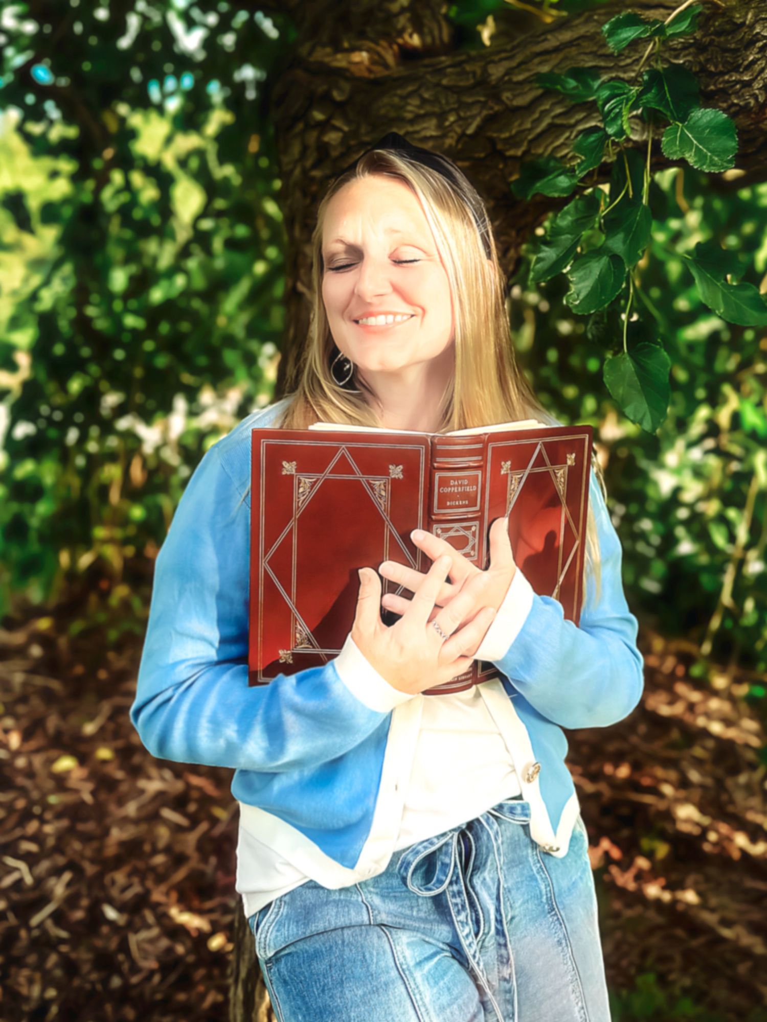 Person holds red book while sitting in lush garden setting wearing denim and blue sweater.