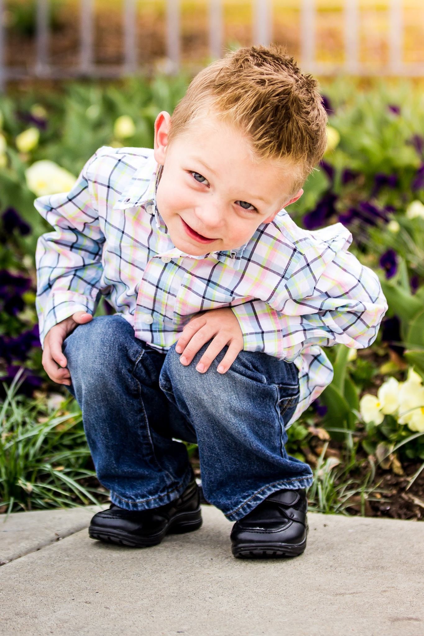Child in plaid shirt crouching by garden flowers during outdoor photo session.