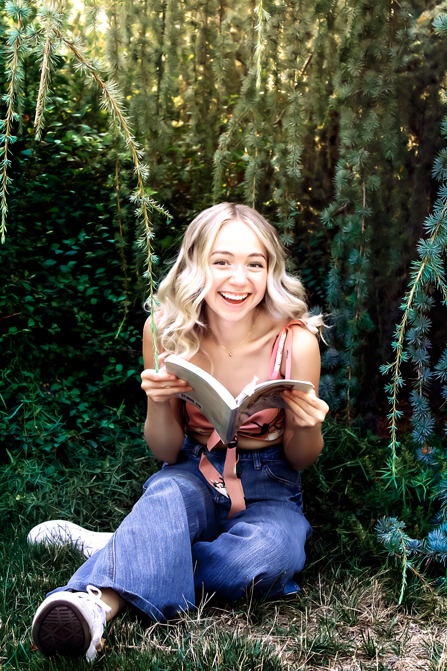 A reader enjoys a book while sitting on grass in a sunlit forest setting wearing jeans and a pink top.