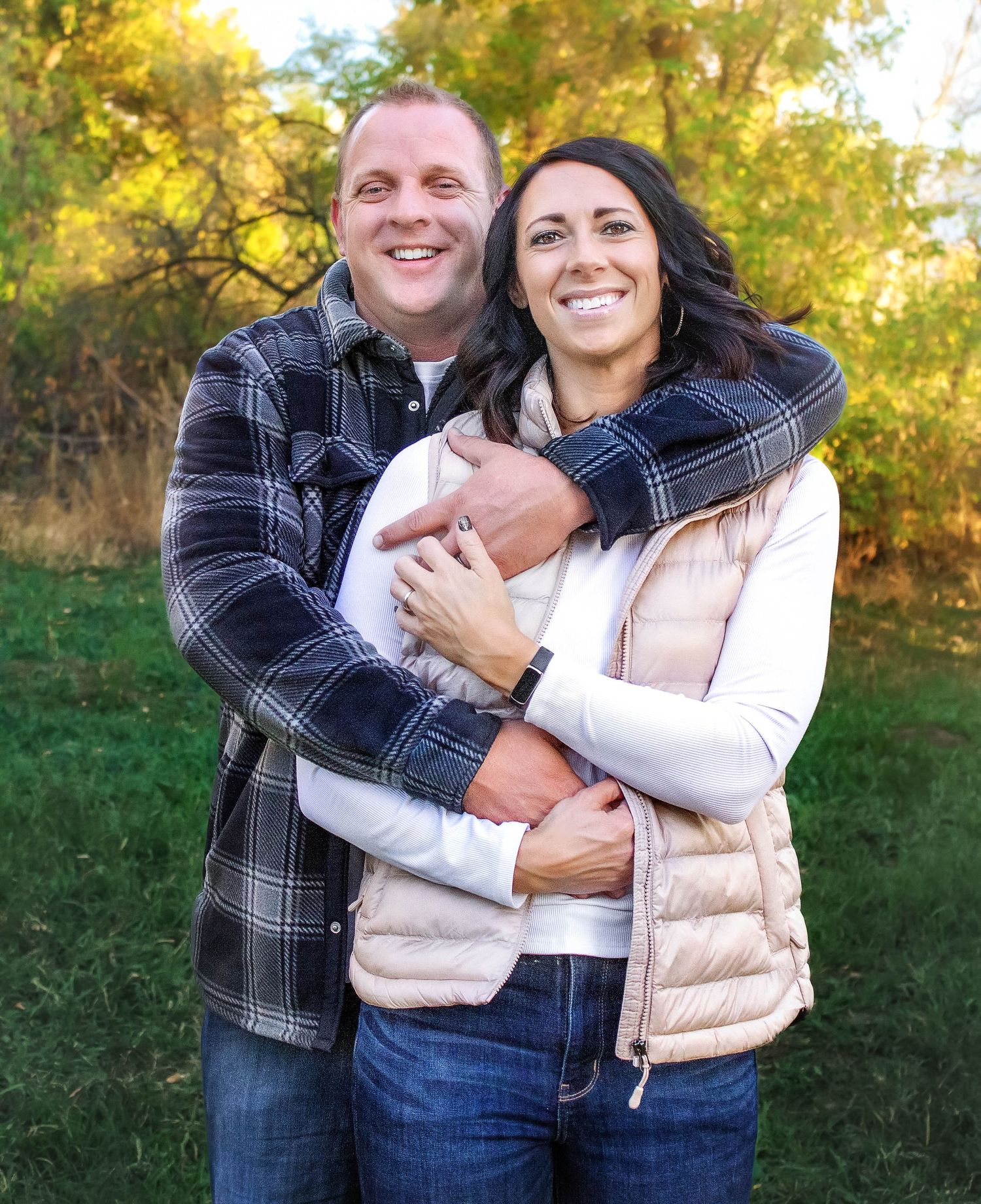A couple embraces outdoors in casual fall attire against a backdrop of golden autumn foliage.