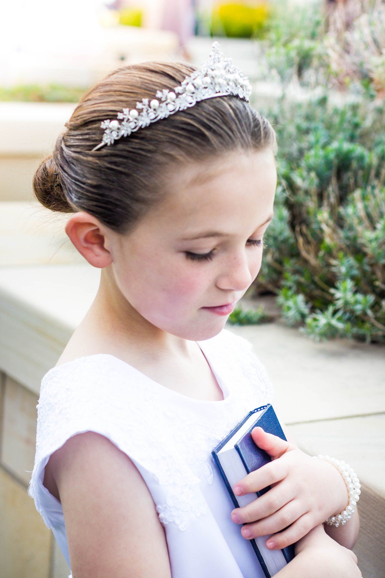 Person in white formal dress wearing tiara poses in garden setting.