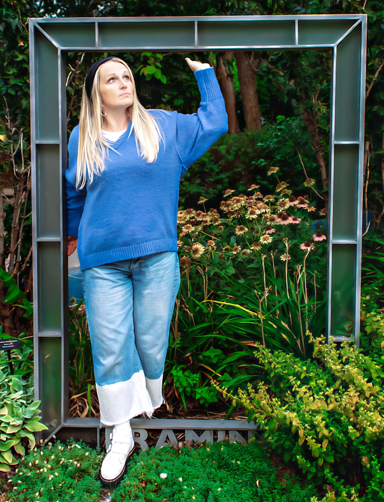 Person in blue sweater and jeans poses near garden arbor and flowering plants.
