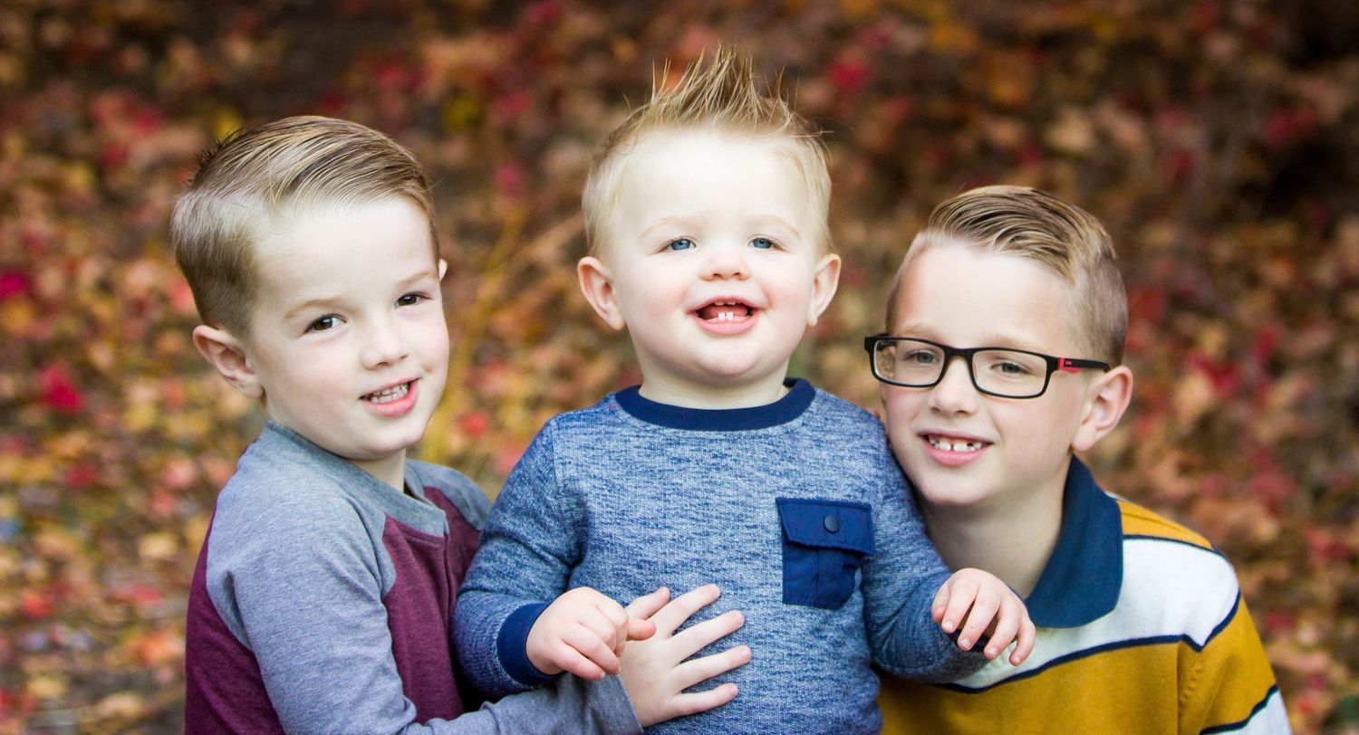 Three young children smile brightly against a background of fall foliage.