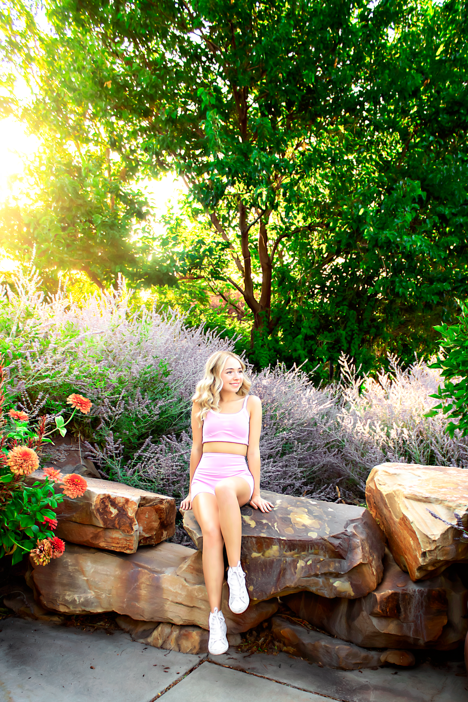 Person in pink athletic wear relaxing on stone wall surrounded by summer garden foliage.