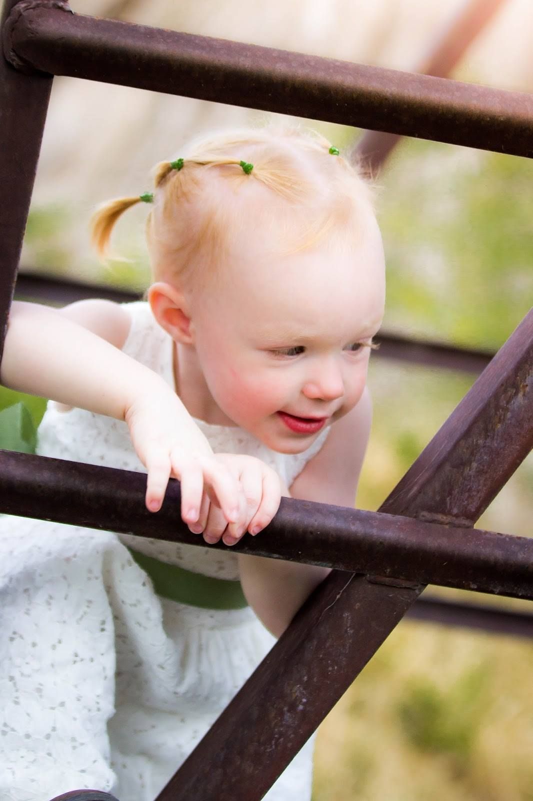 Child in white dress peeks through metal railing with joyful expression.