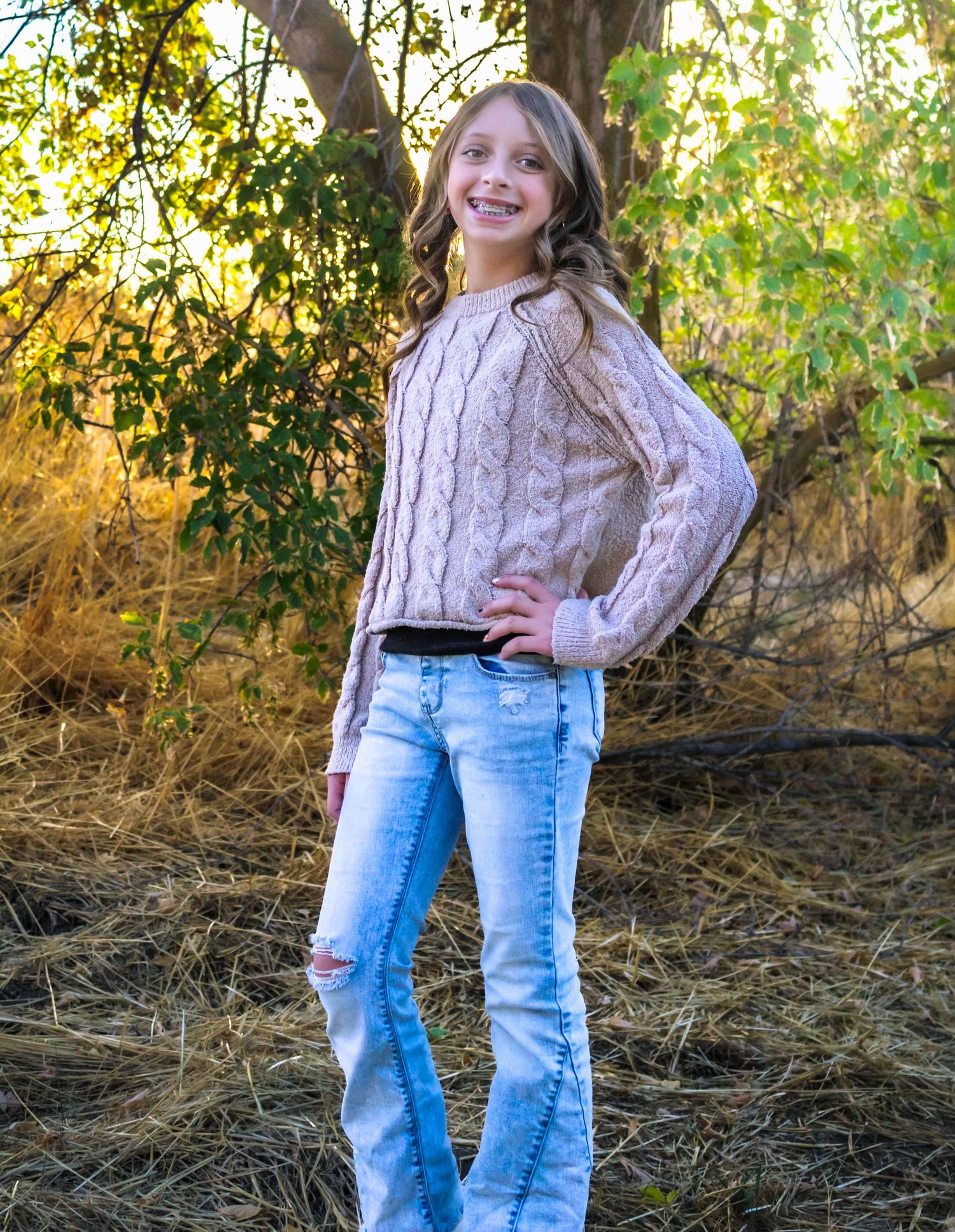 A child in cable knit sweater and jeans poses playfully outdoors in autumn setting.