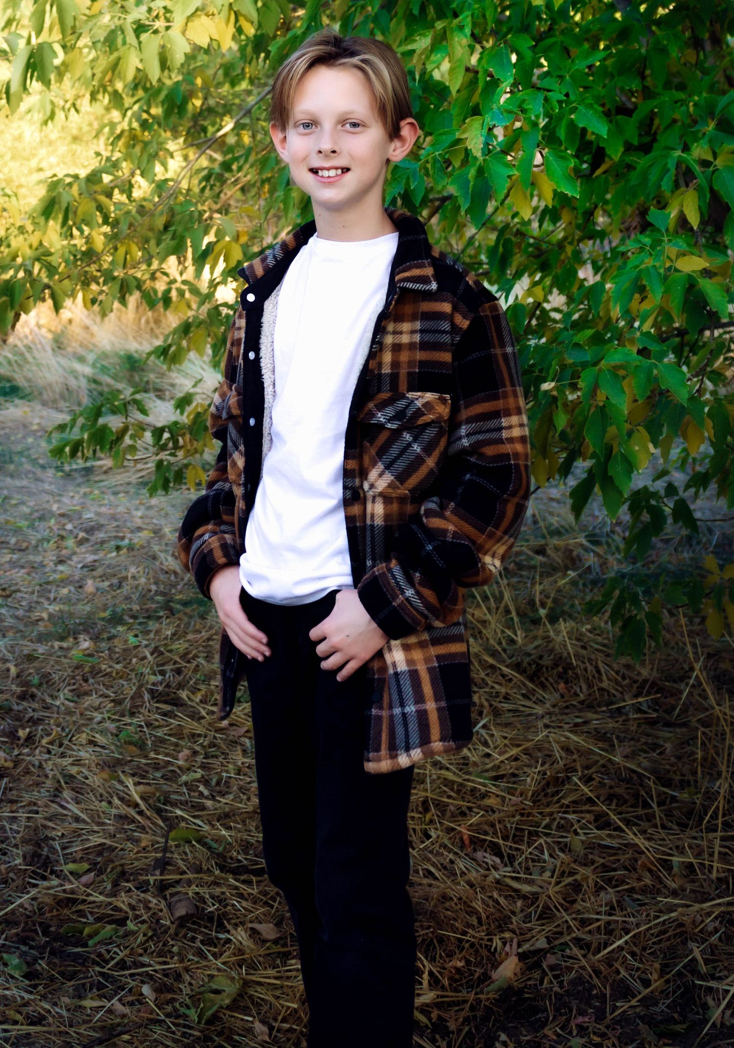 A child poses in plaid flannel and dark pants against lush green foliage.