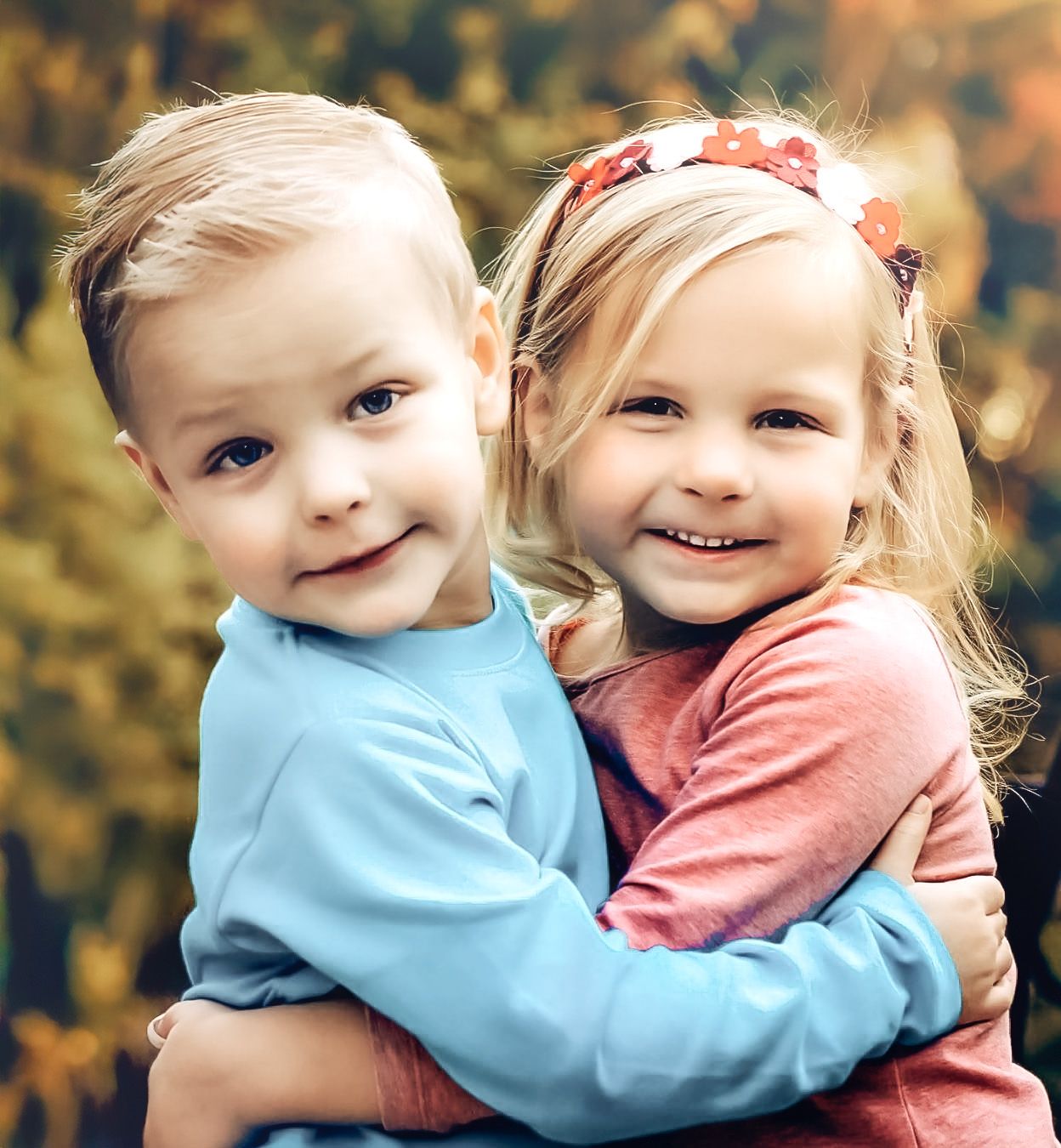 Two children in blue and pink shirts hugging and smiling together in an outdoor setting.