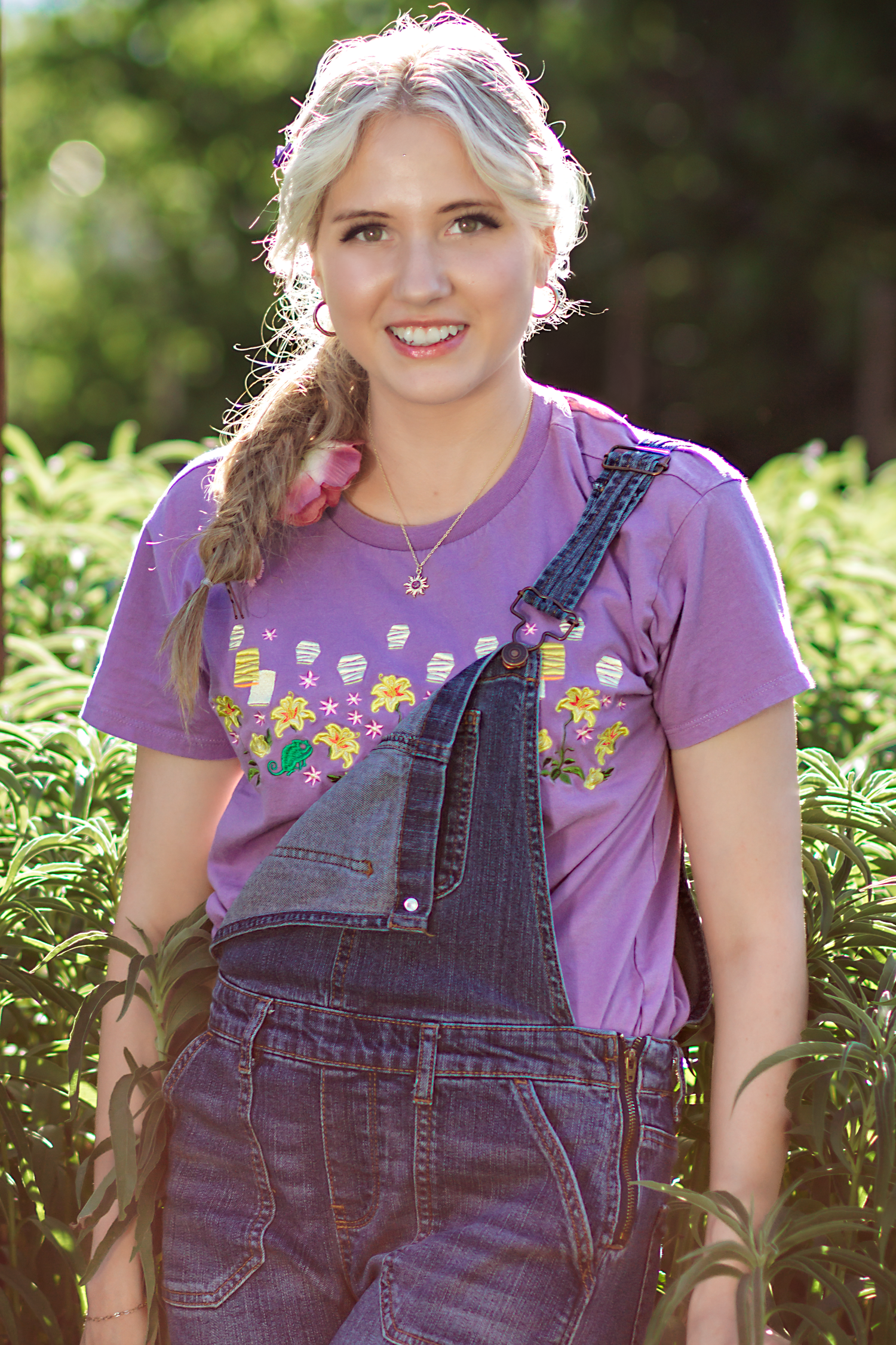 Person in denim overalls and purple floral shirt posing in a sunny garden.