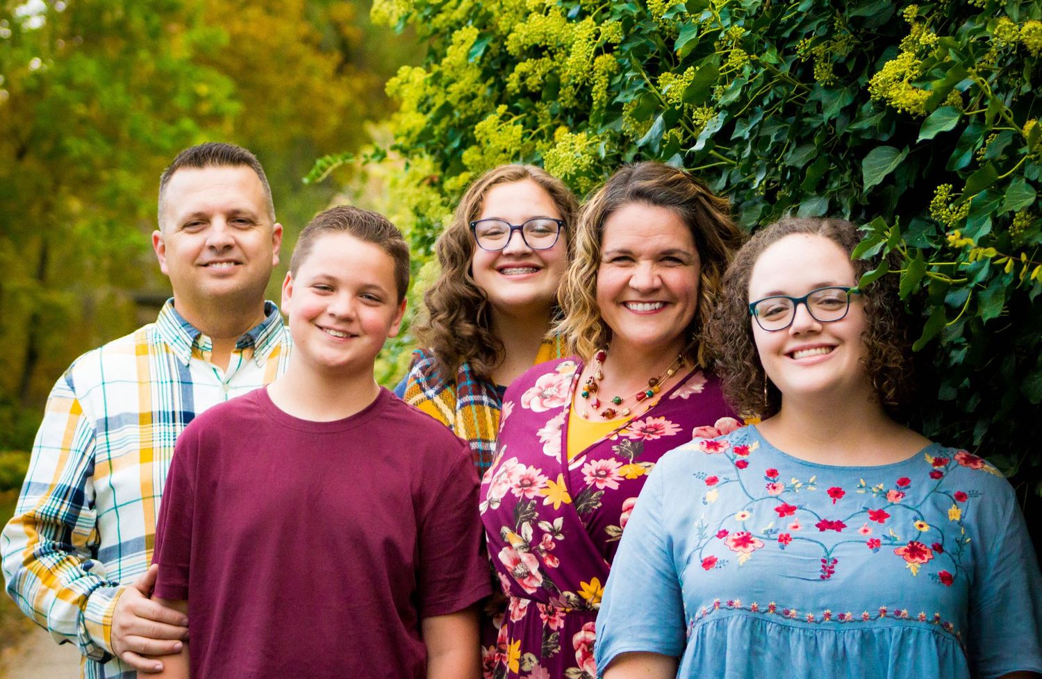 A family of five smiles together during an outdoor portrait session with green foliage.