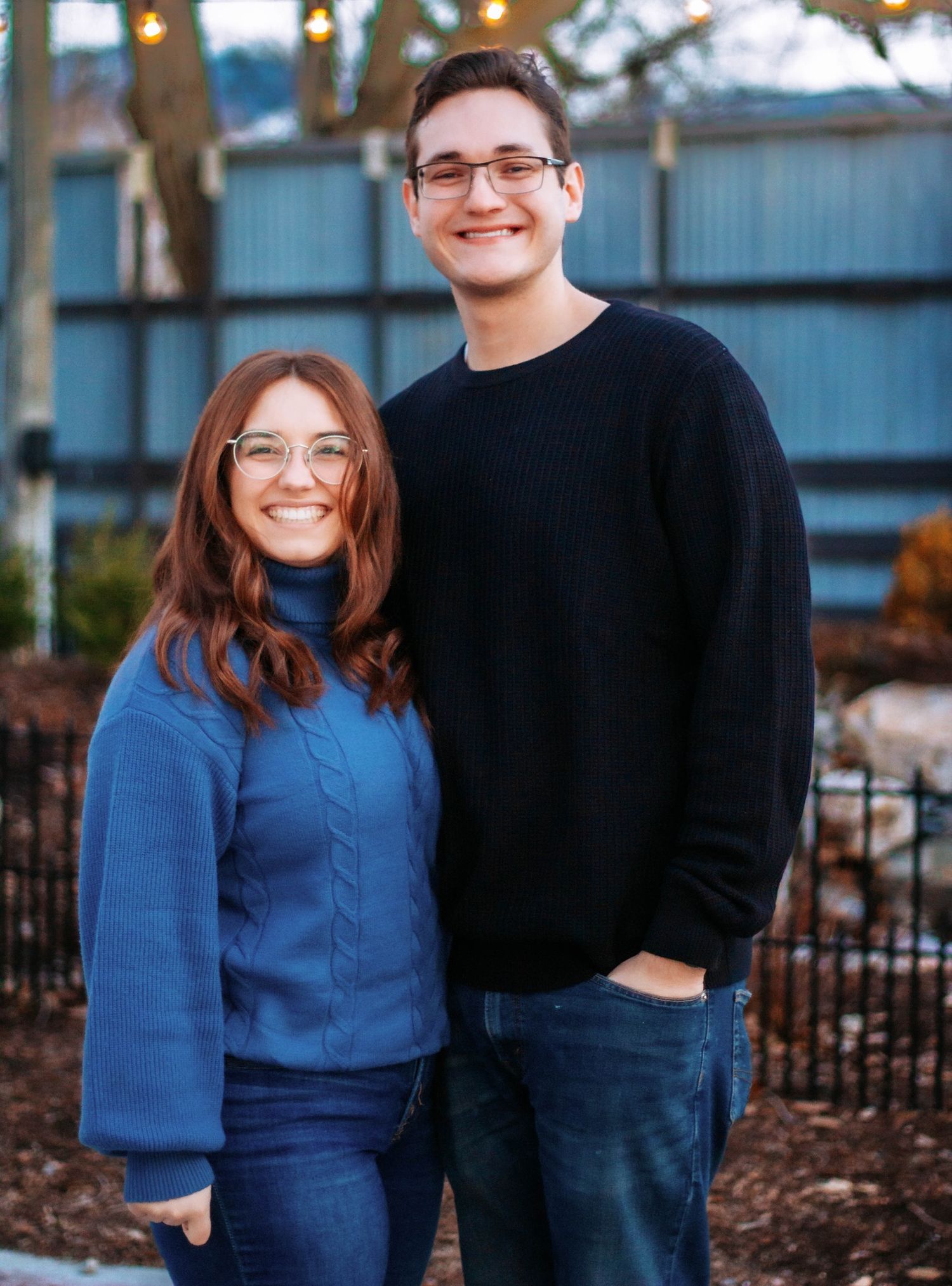 Couple wearing coordinating blue and black outfits pose together in front of an outdoor autumn backdrop.