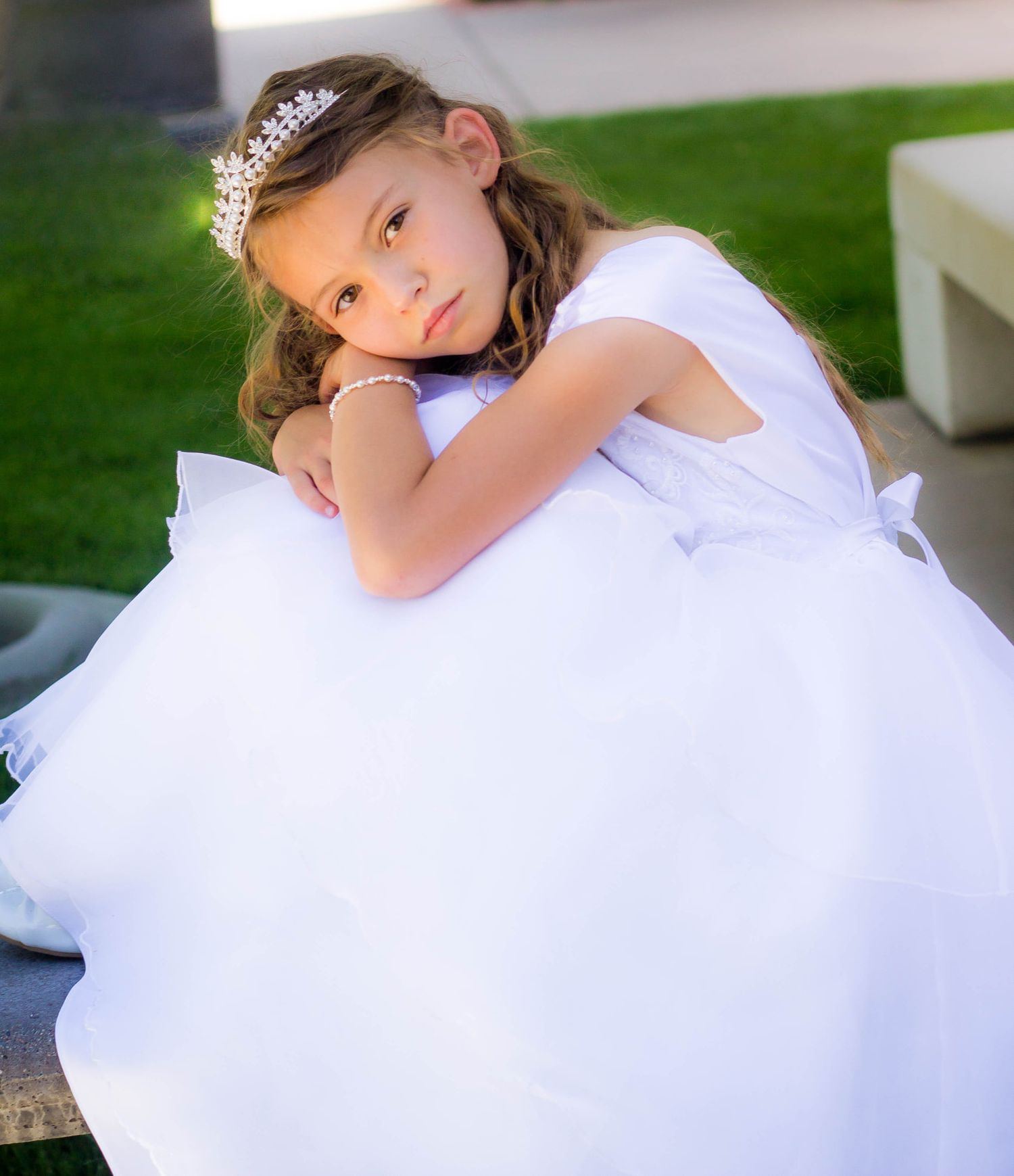Child in white dress poses gracefully on outdoor furniture.