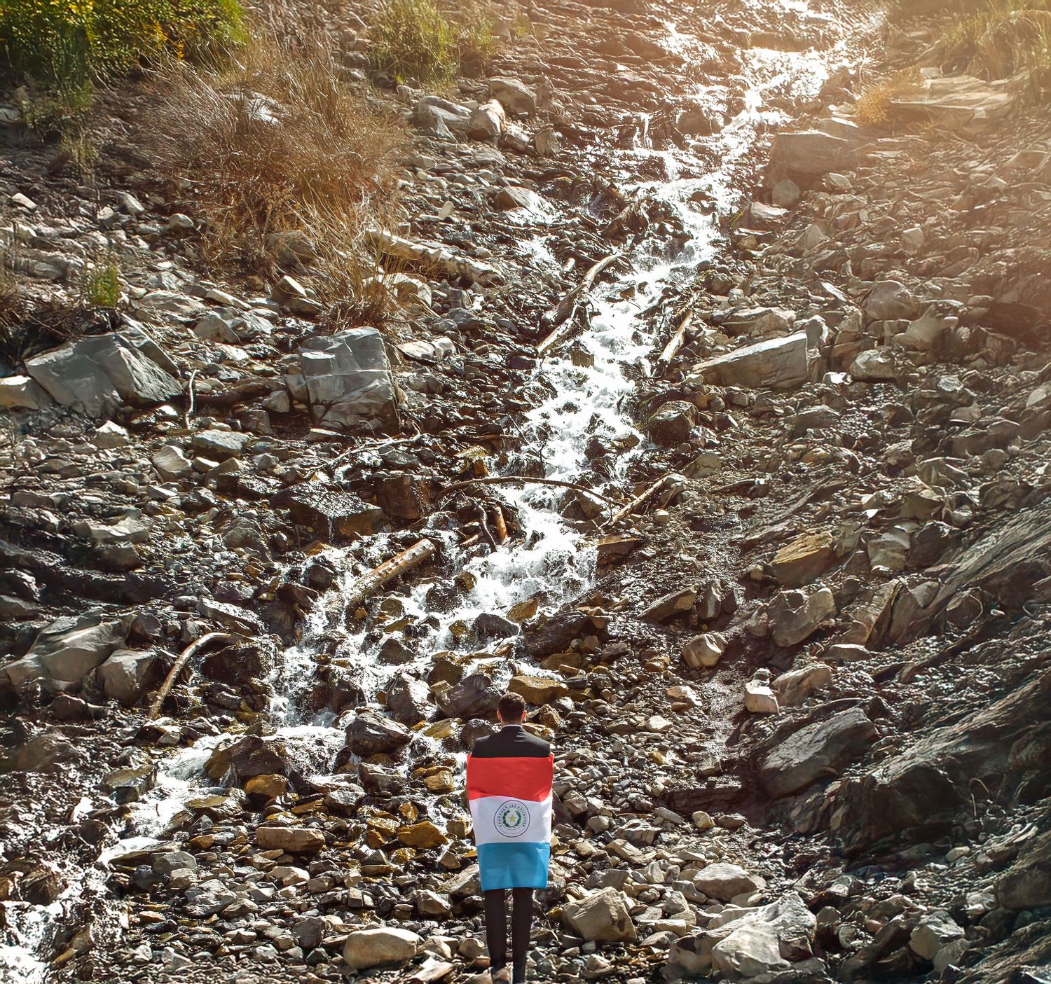 A small Paraguay flag stands near a rocky waterfall stream in a mountainous landscape.