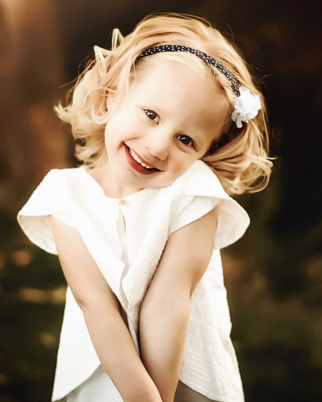 Child in white dress smiles playfully against dark background.
