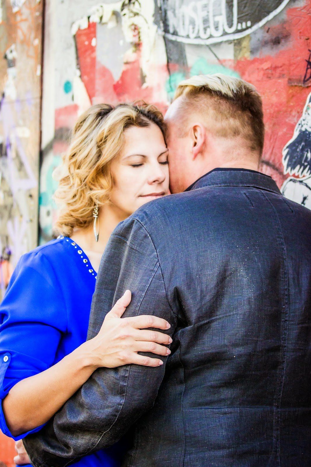 Couple in blue dress and dark suit pose against colorful graffiti wall.